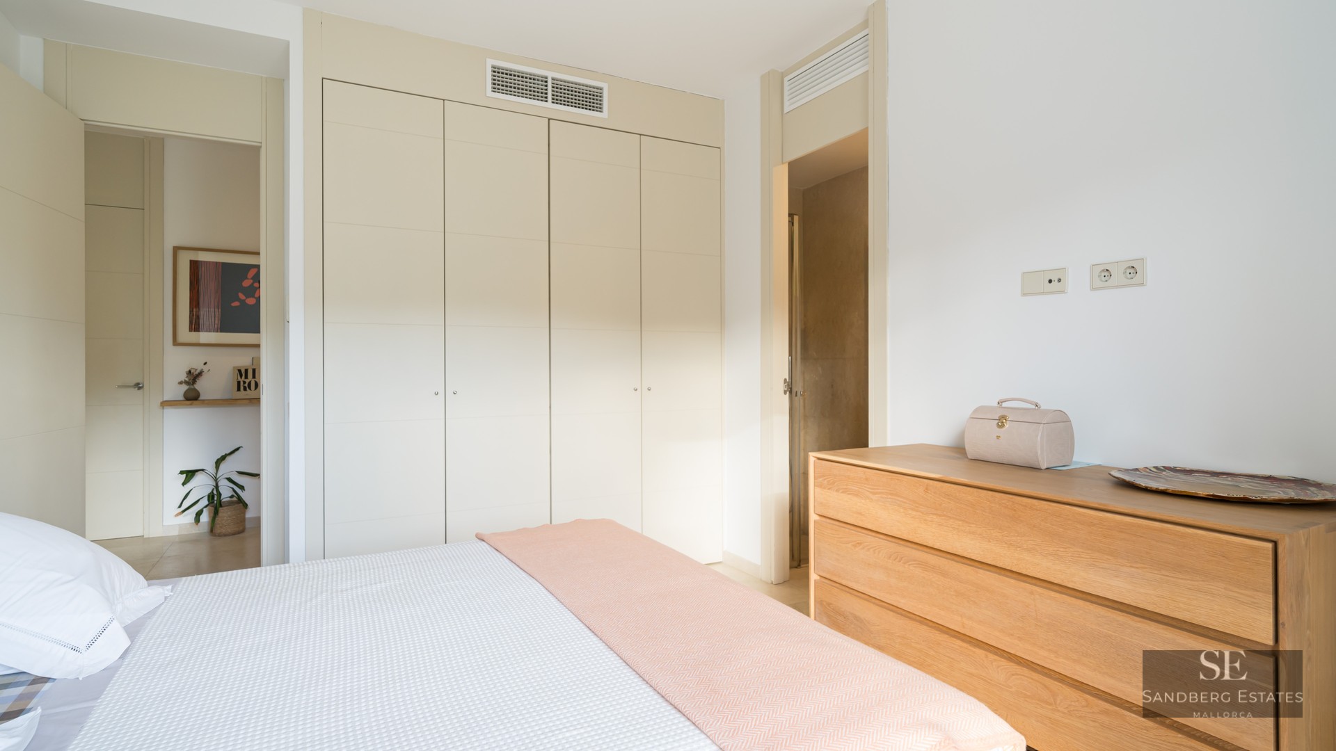 Bright bedroom featuring cream built-in wardrobes, a wooden chest of drawers, and a bed with a pastel pink throw.