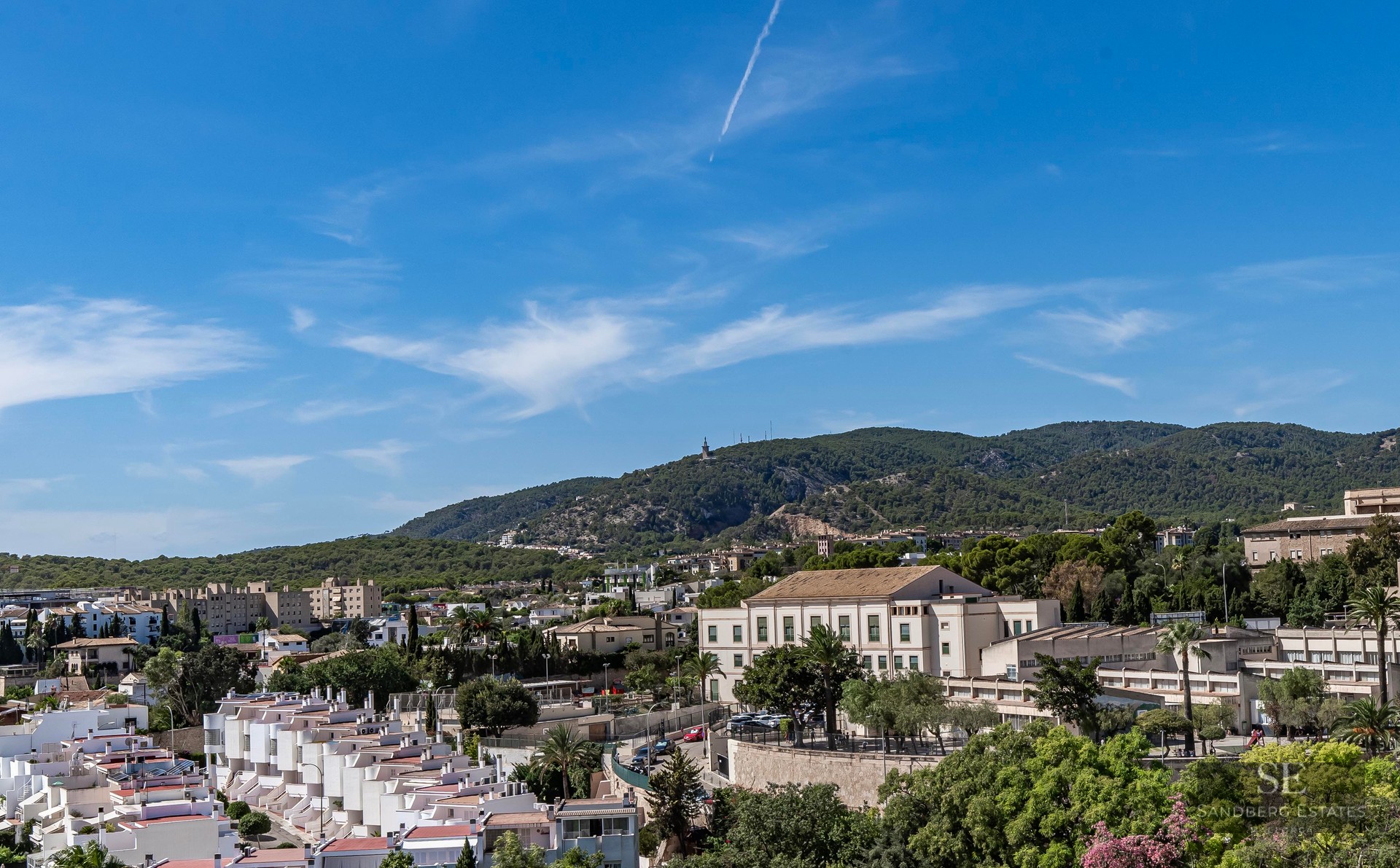 High-angle view of white houses in a Mediterranean town with lush green mountains and a blue sky in the background.
