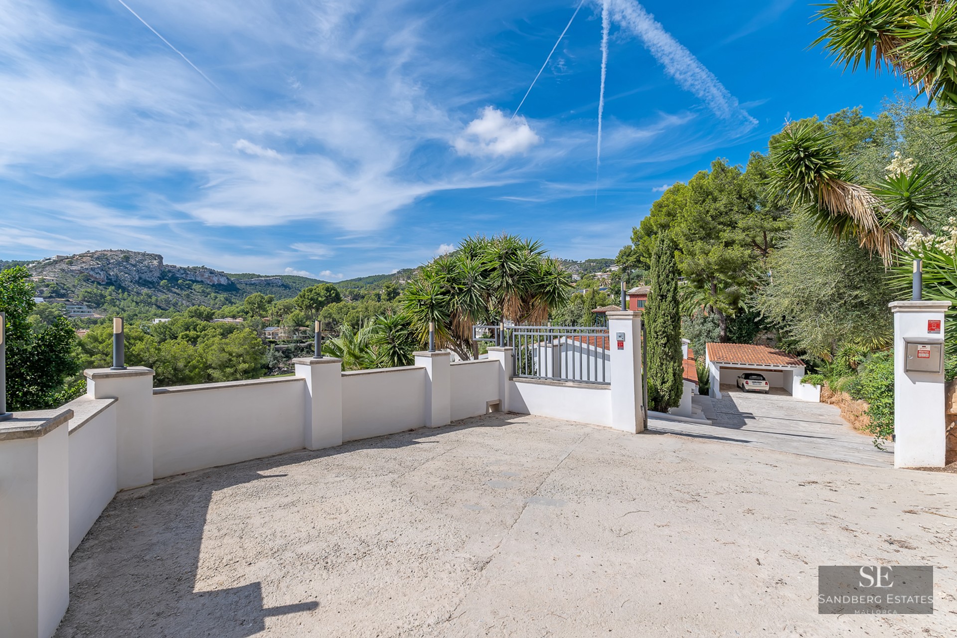 A wide concrete driveway surrounded by white walls and palm trees, leading to a garage with mountain views in the distance.