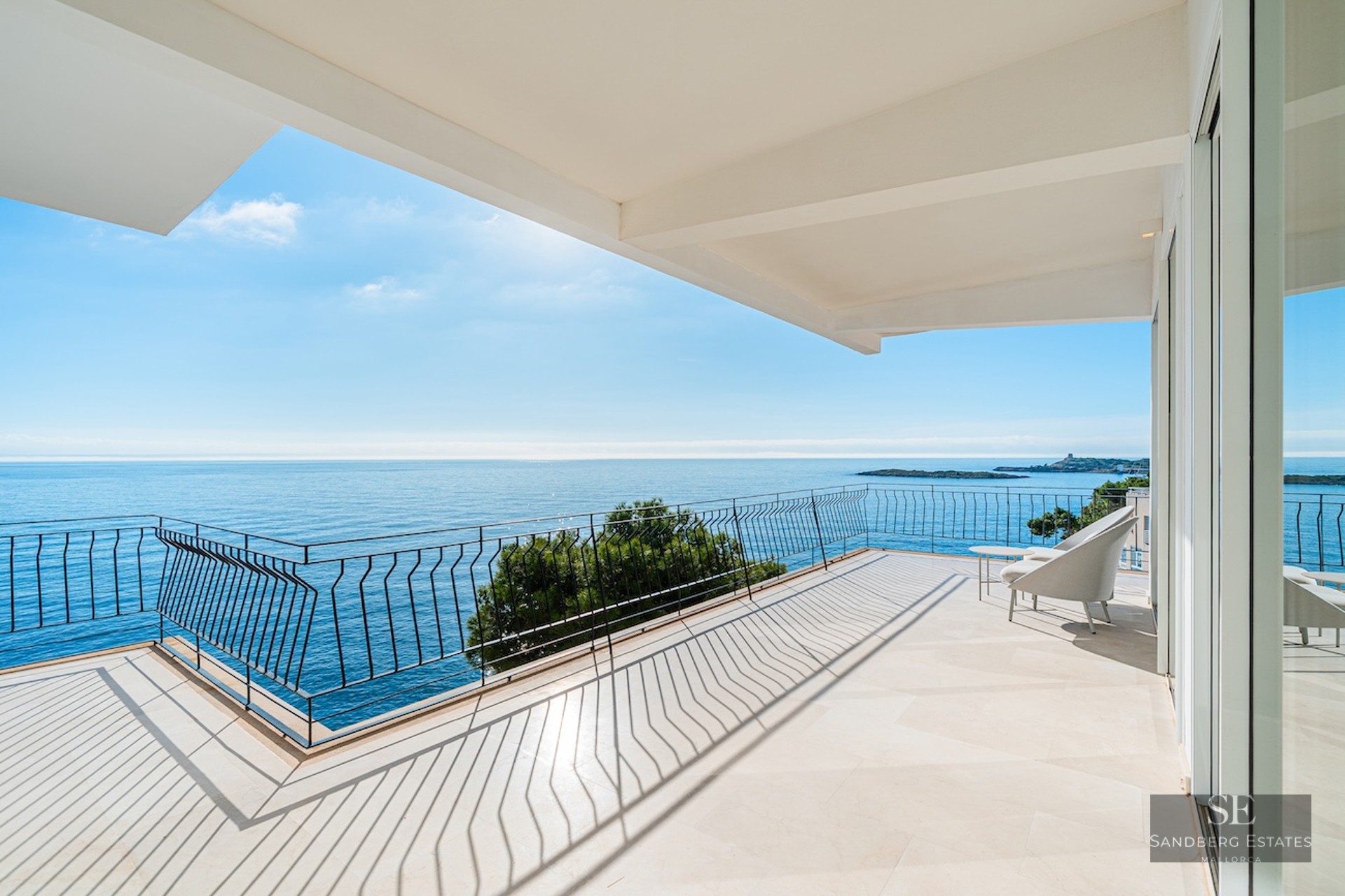 Wide modern terrace with stone flooring and black railing overlooking a calm blue sea under a bright sky.