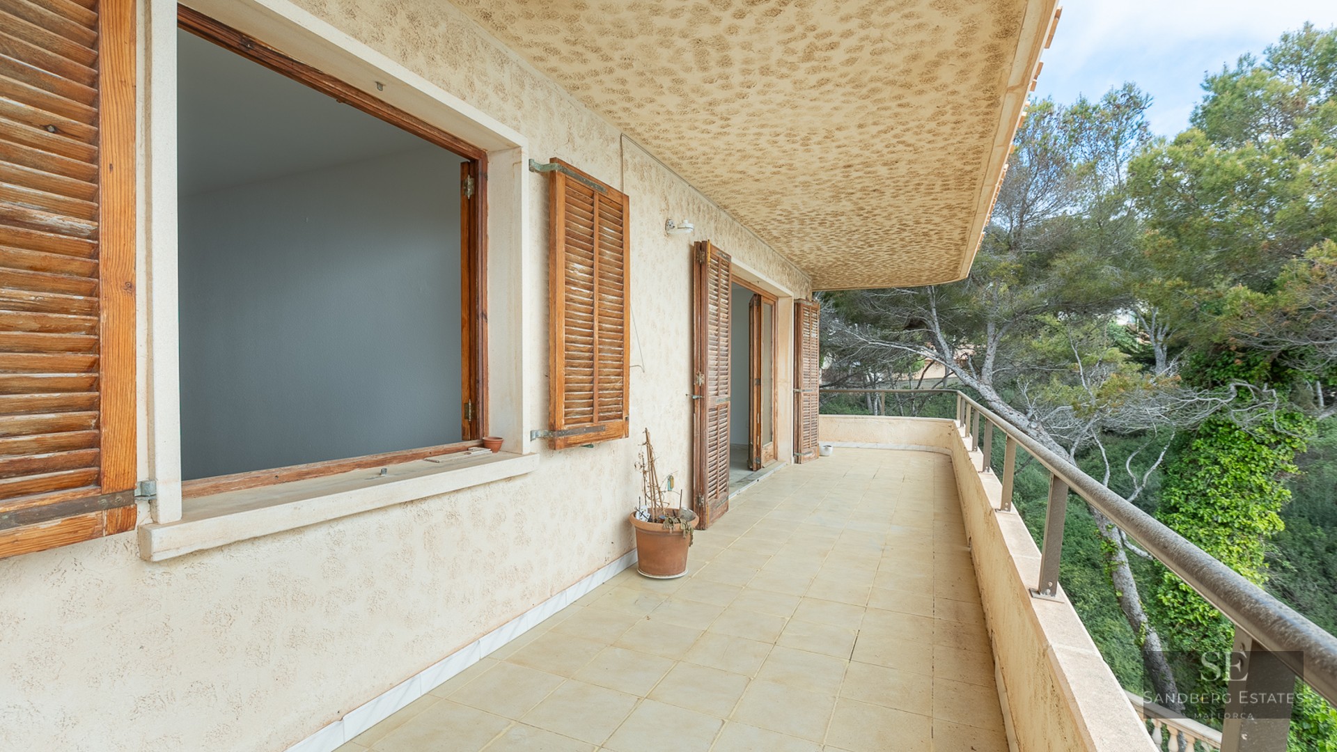 A long balcony with beige tiles, wooden shutters, and a metal railing overlooking lush green trees.