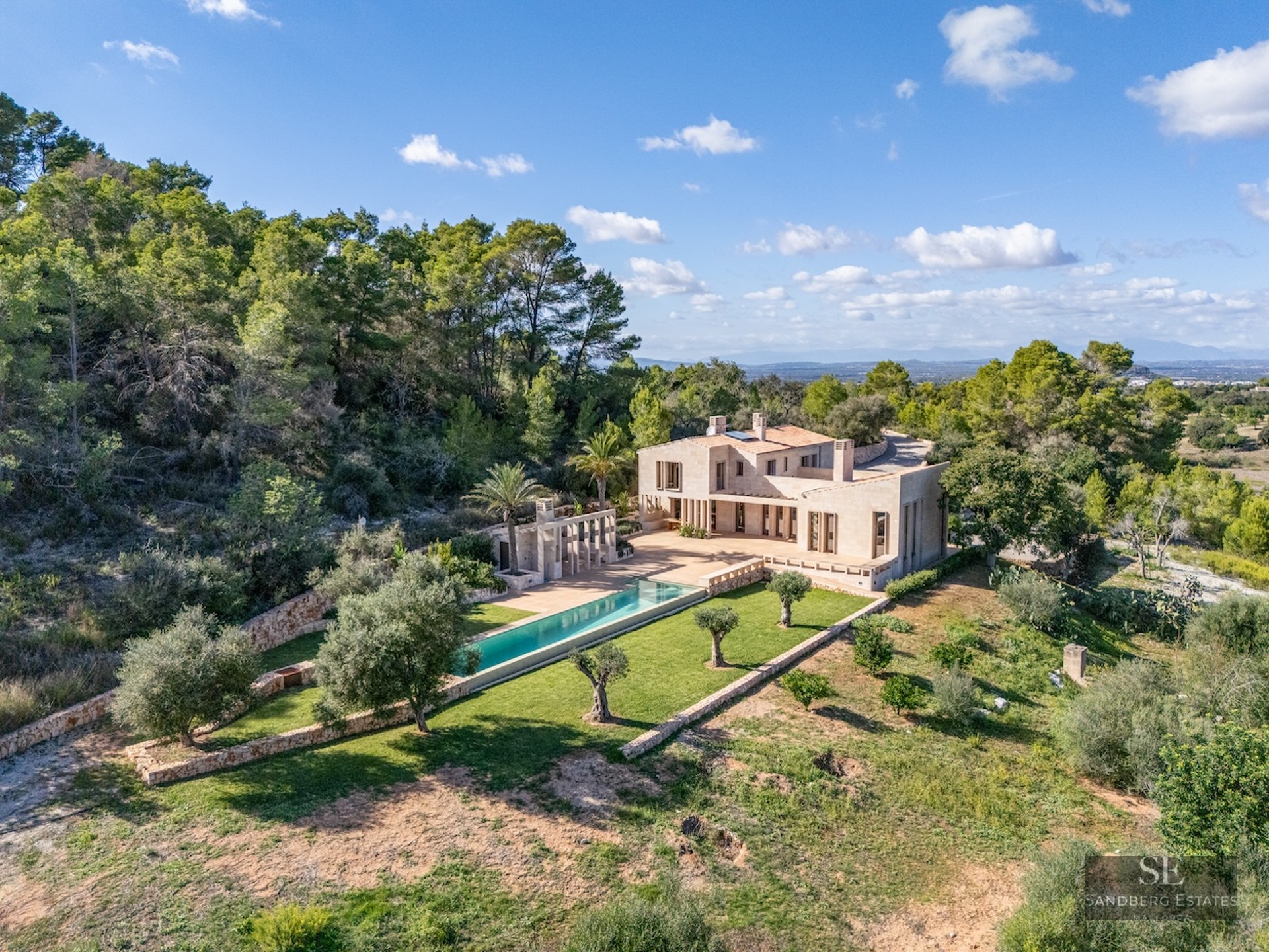 Aerial view of a luxury stone villa with a long turquoise pool surrounded by olive trees and lush forest.