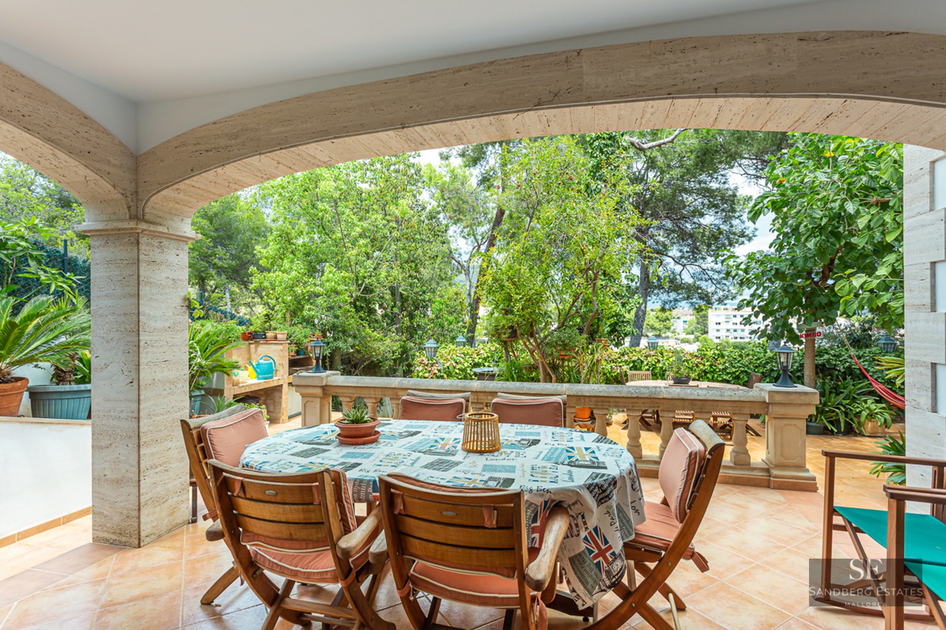 A covered stone terrace with a round dining table, wooden chairs, and a stone balustrade overlooking a lush garden.