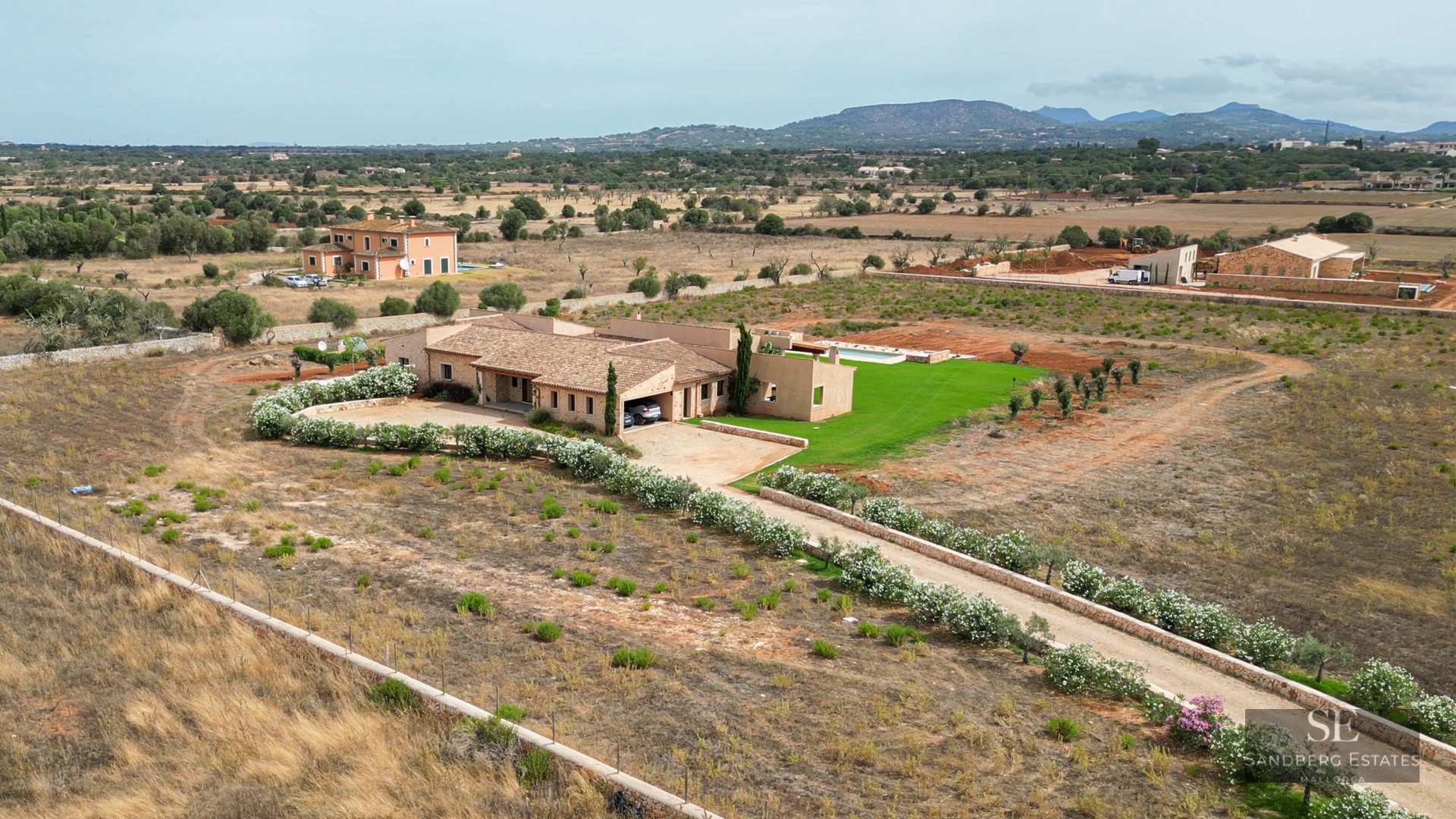 Aerial shot of a stone villa with a pool and flower-lined driveway in a Mediterranean countryside setting.