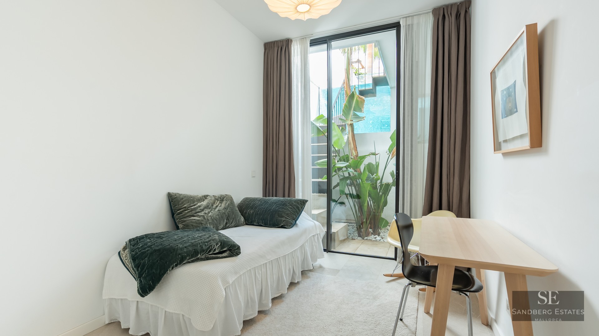 Bright bedroom with a single bed, wooden desk, and glass door opening to a small outdoor garden with plants.