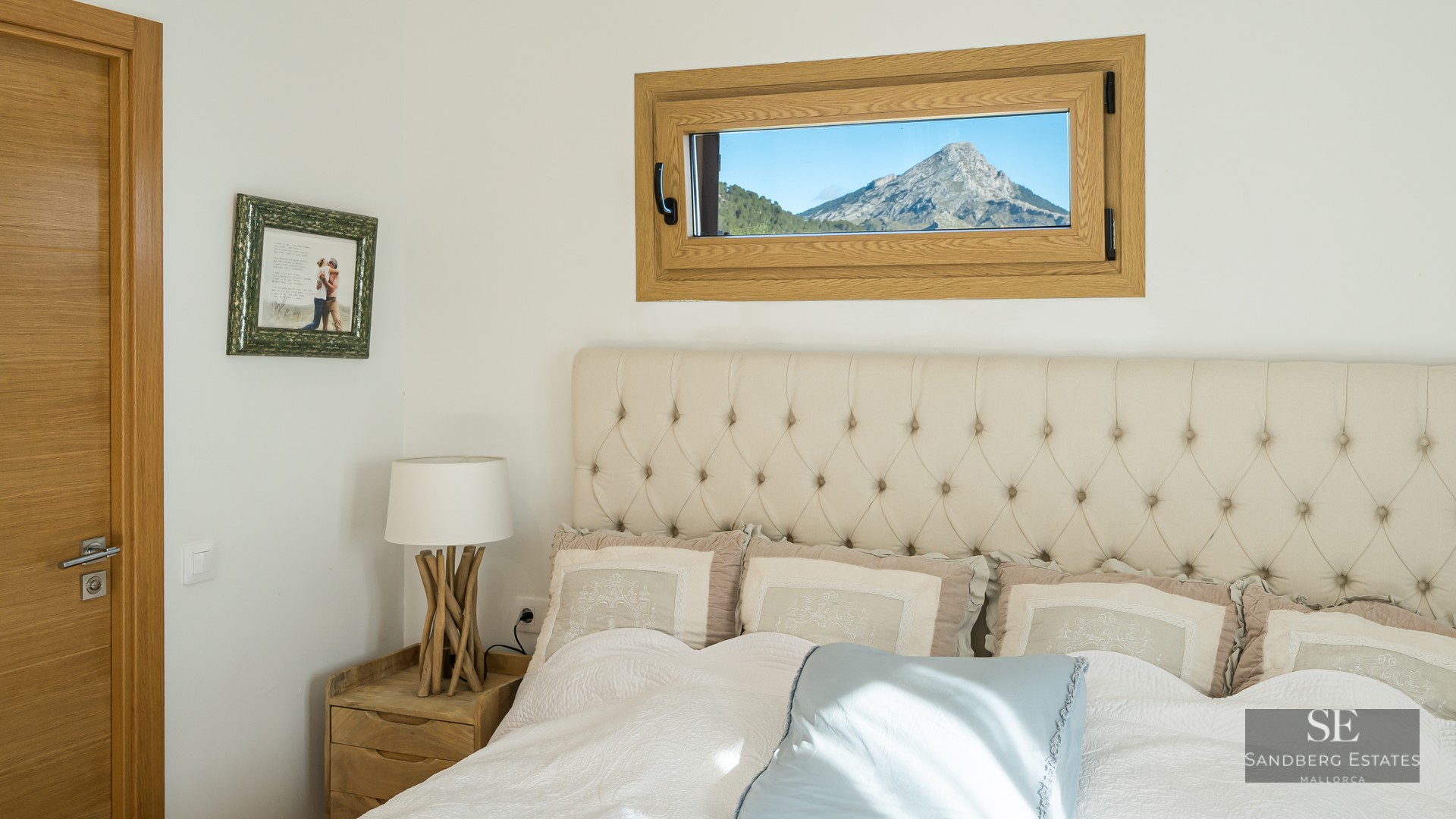 Bed with beige tufted headboard next to a small horizontal window framing a rugged mountain peak.