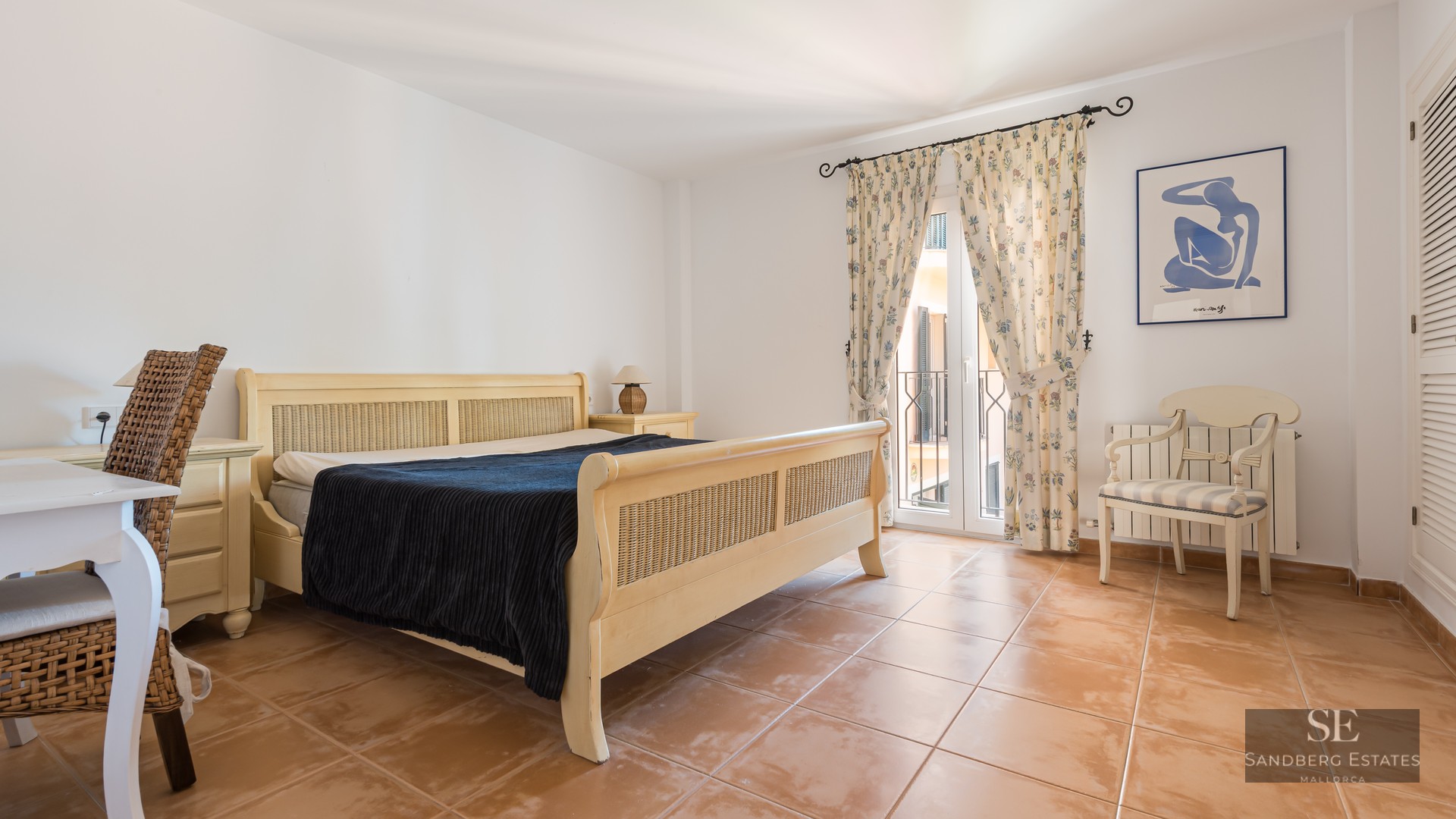 Bedroom with wicker bed frame, terracotta tile floor, desk, and balcony with floral curtains.