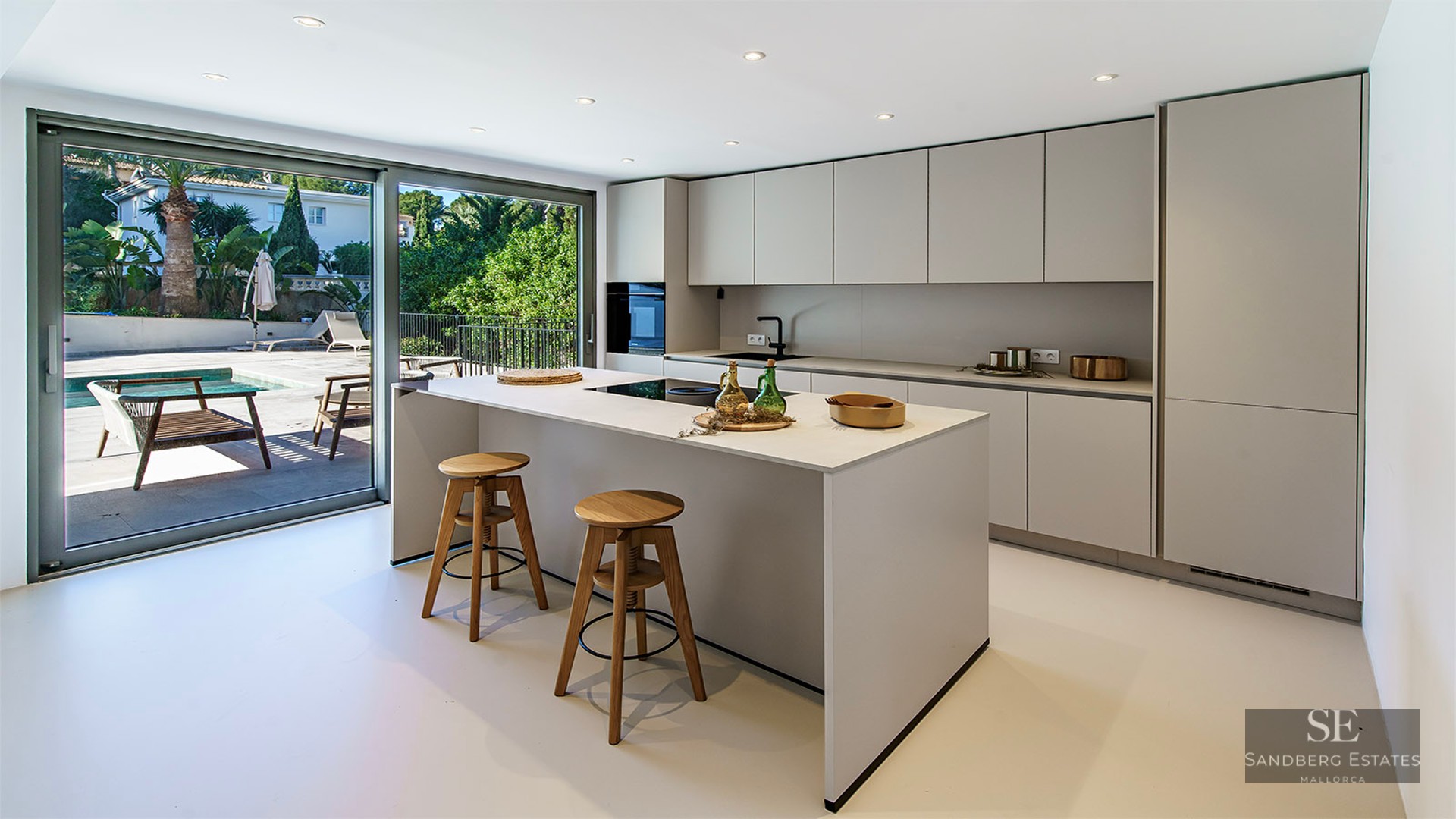 Modern white kitchen with central island, two wooden stools, and large glass sliding doors opening to a terrace and pool.