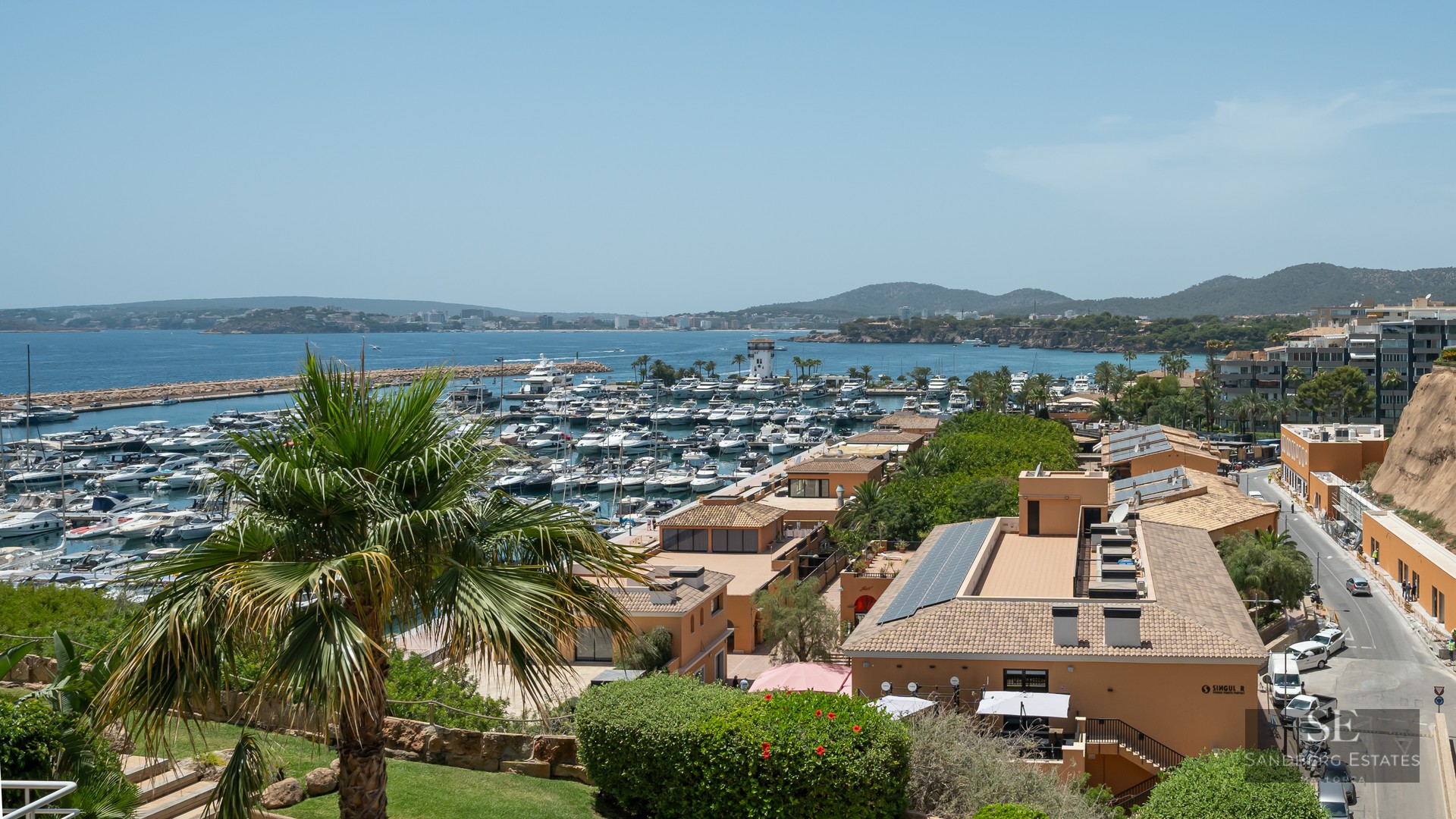 Elevated view of a Mediterranean marina filled with luxury yachts, overlooking the sea and coastline under a clear sky.