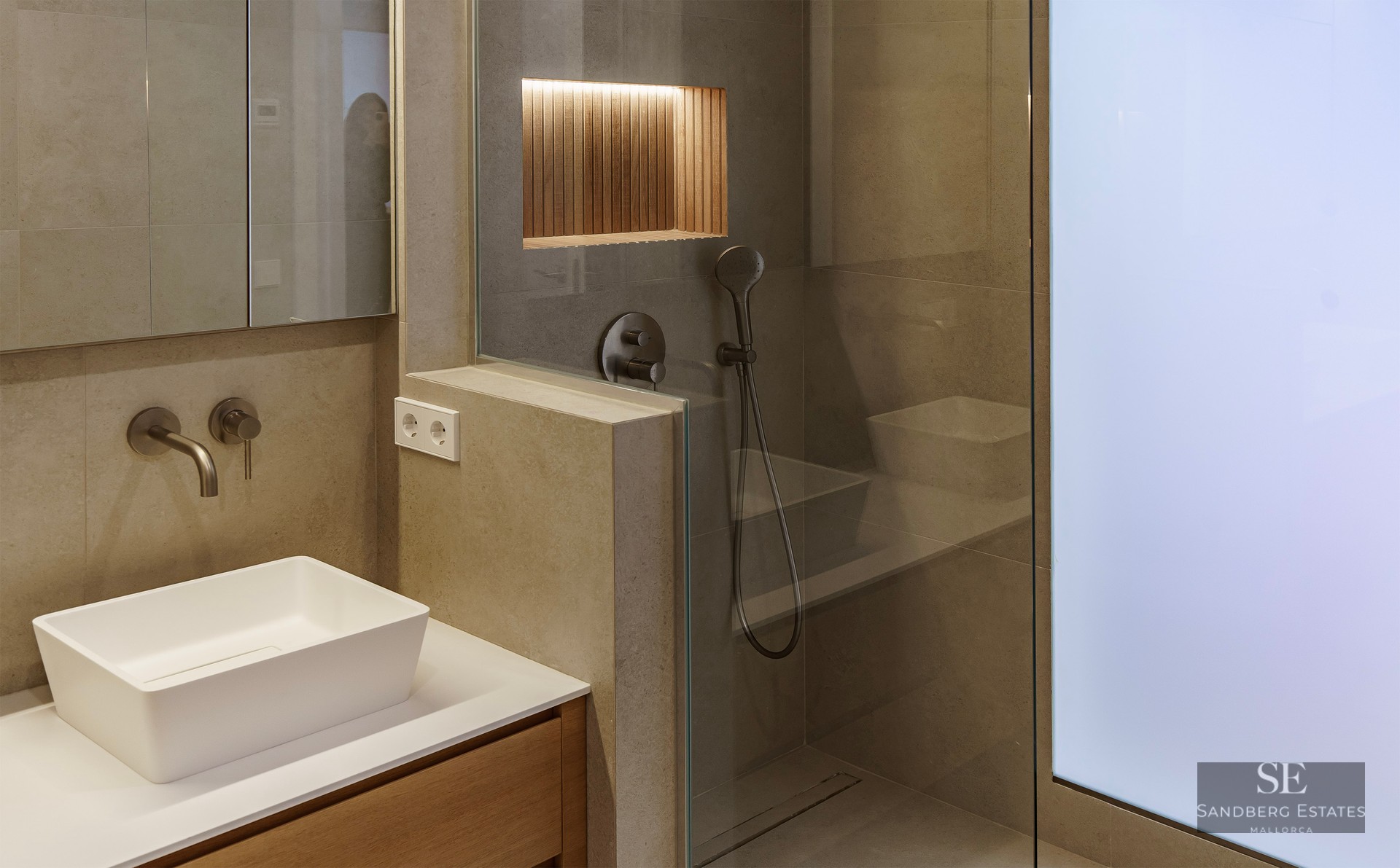 Salle de bain moderne avec vasque à poser, murs en pierre et douche vitrée avec niche en bois éclairée.