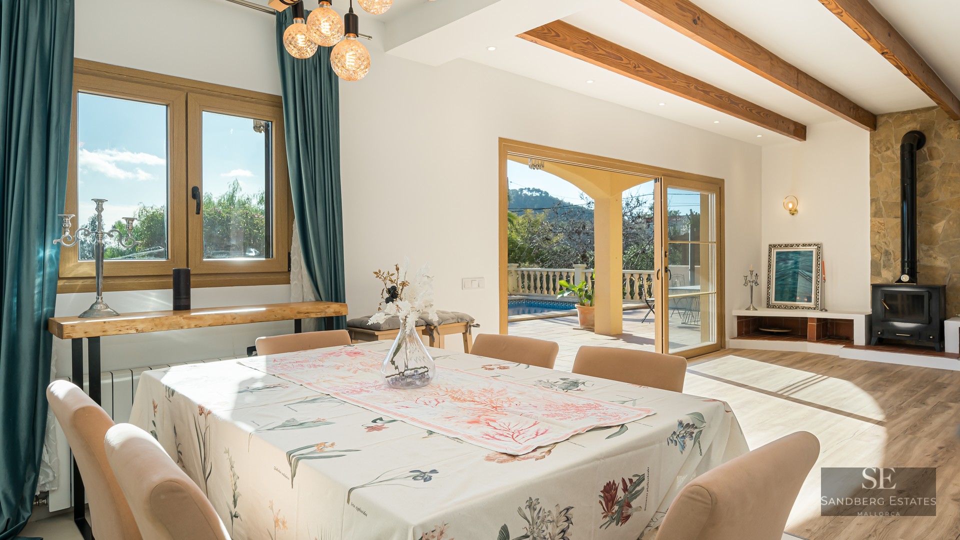 Bright dining room with wooden beams, stone fireplace, and glass doors leading to a terrace with a swimming pool.