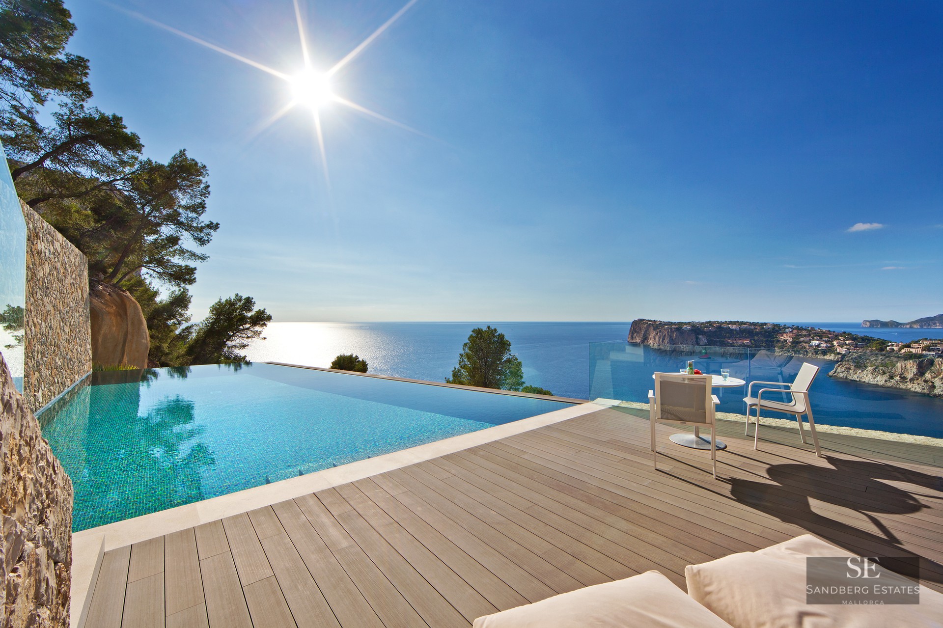 Infinity pool with turquoise water on a wooden deck overlooking a blue sea and rugged coastline under a bright sun.