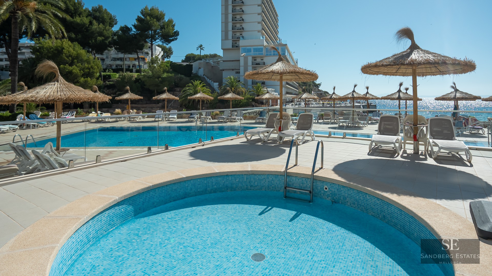 Bright outdoor pool area with straw umbrellas, white sun loungers, glass railings, and a view of the sea.