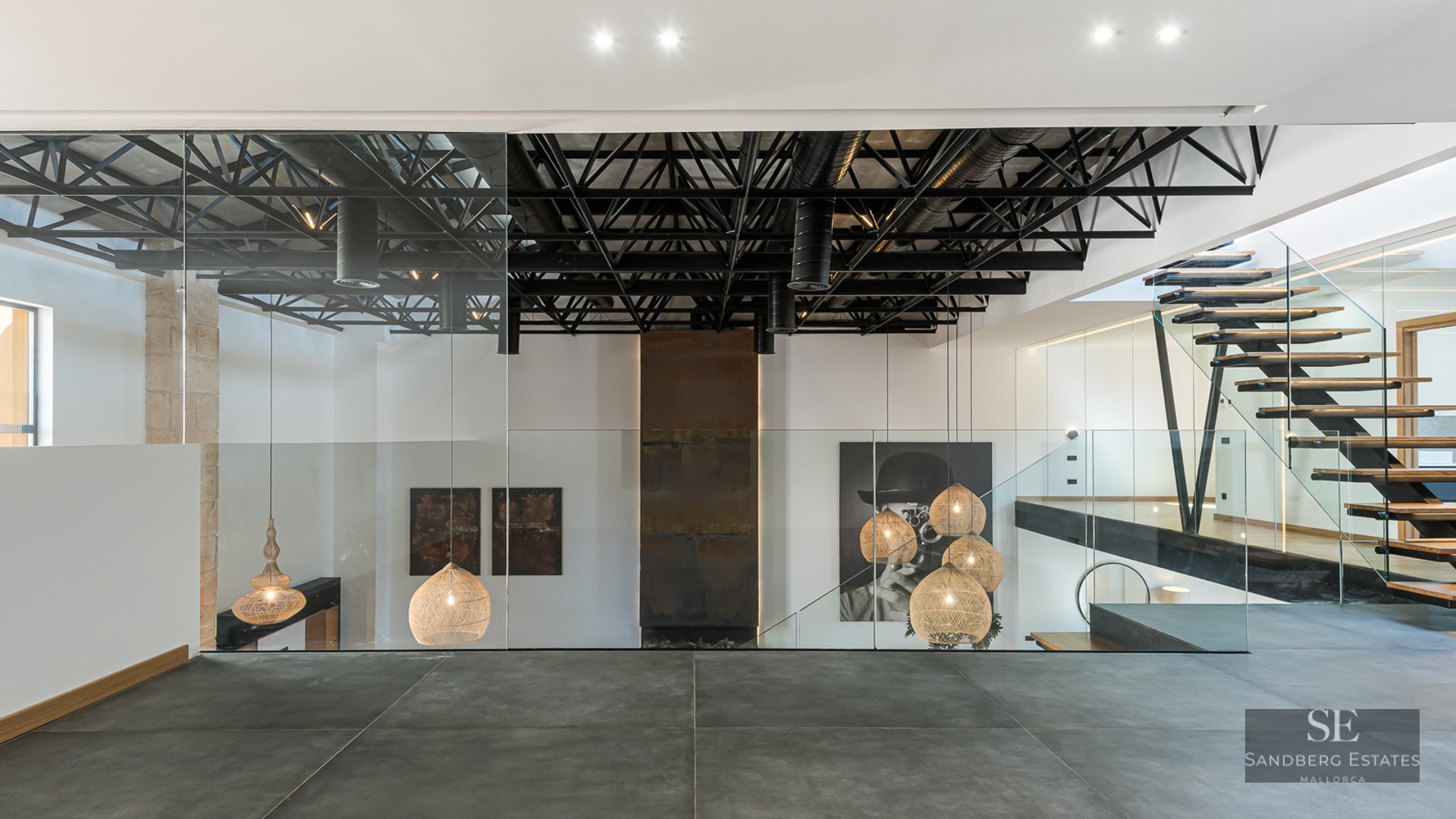 Modern interior featuring black steel ceiling trusses, glass railings, floating wood stairs, and designer pendant lamps.