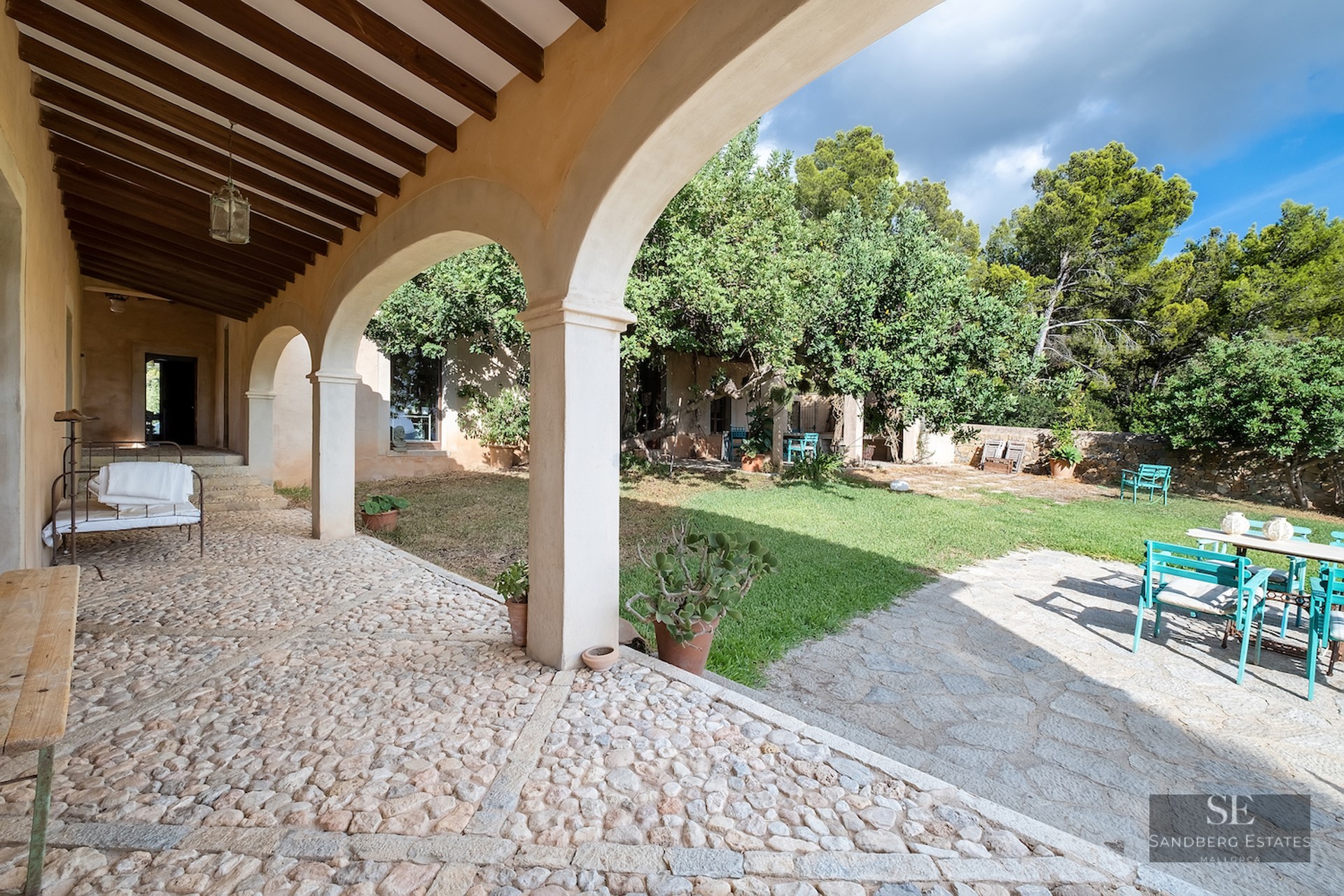 A traditional stone-arched porch with cobblestone flooring looking out onto a sunny garden with turquoise seating.