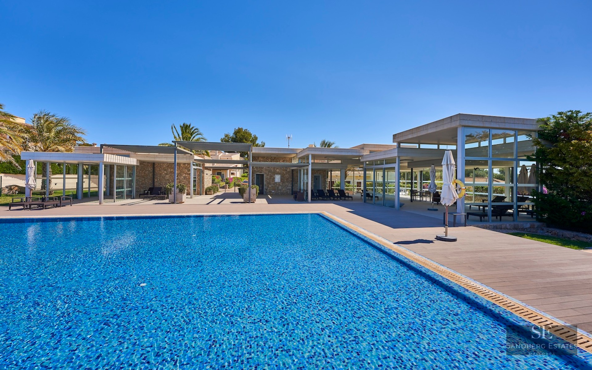 Large rectangular swimming pool with blue tiles next to a modern stone and glass villa under a clear blue sky.