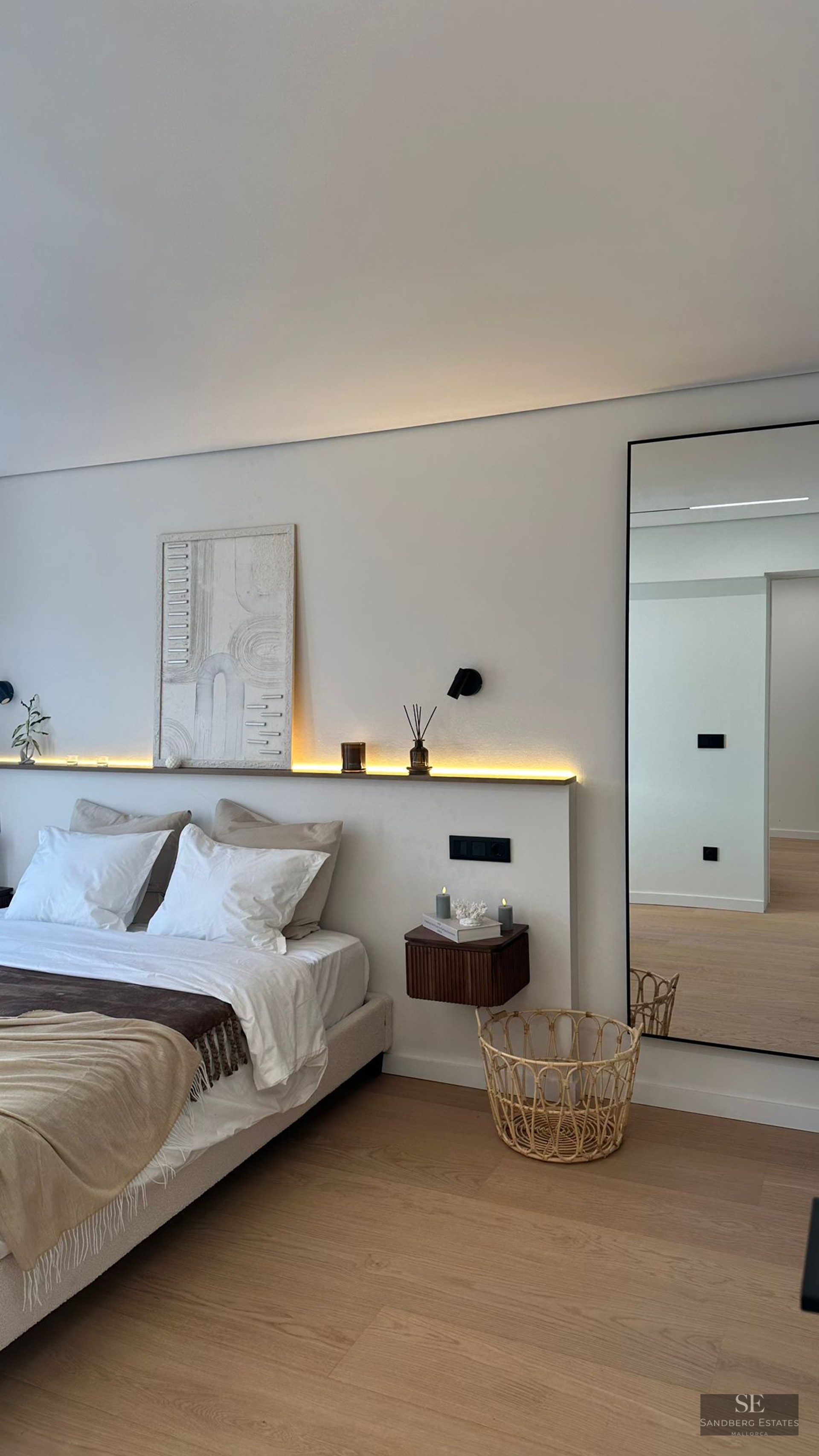 Modern bedroom featuring a white bed, LED backlighting, textured art, and a large mirror on light wood floors.
