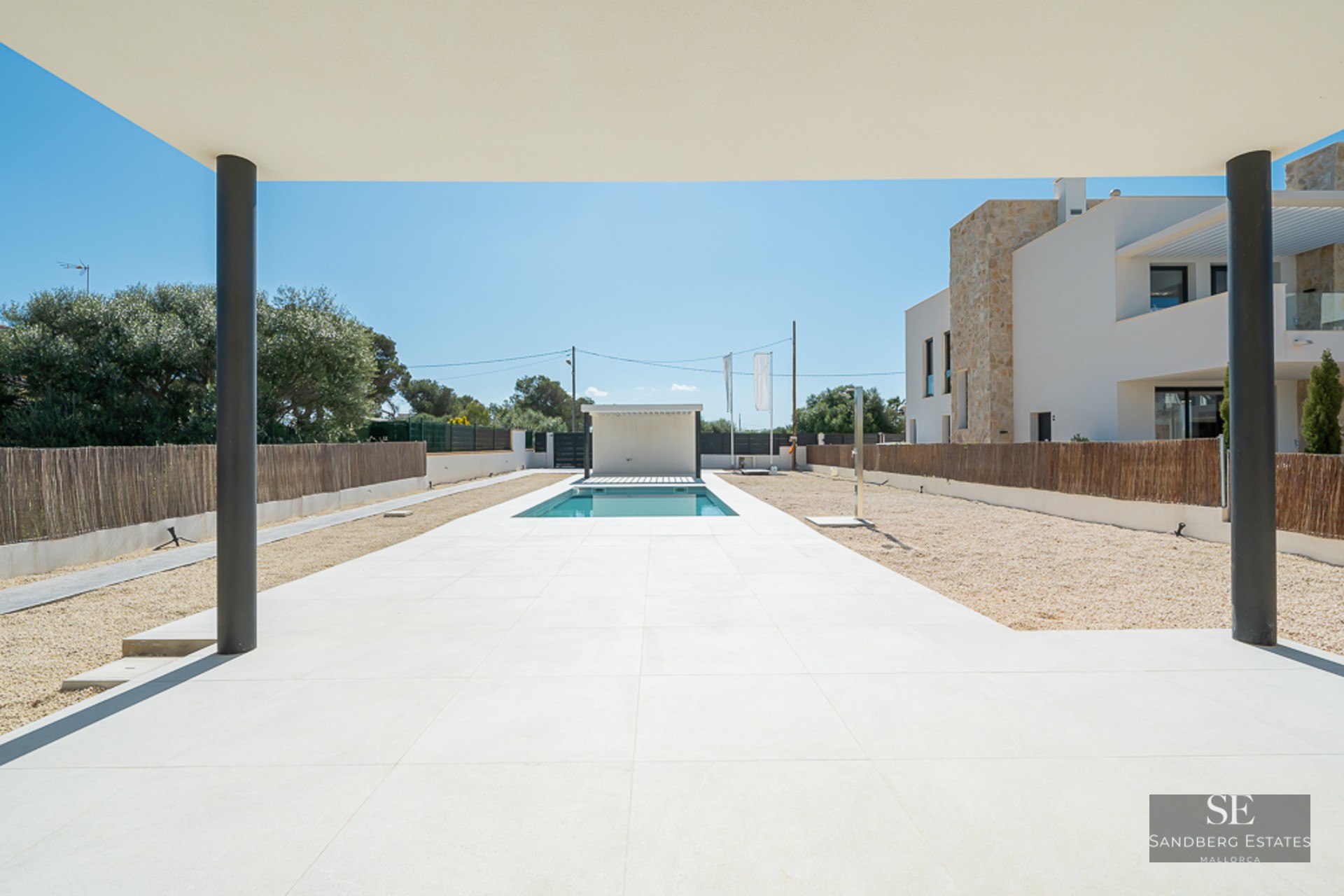 View from a covered porch toward a rectangular pool surrounded by white tiles and gravel in a modern villa setting.