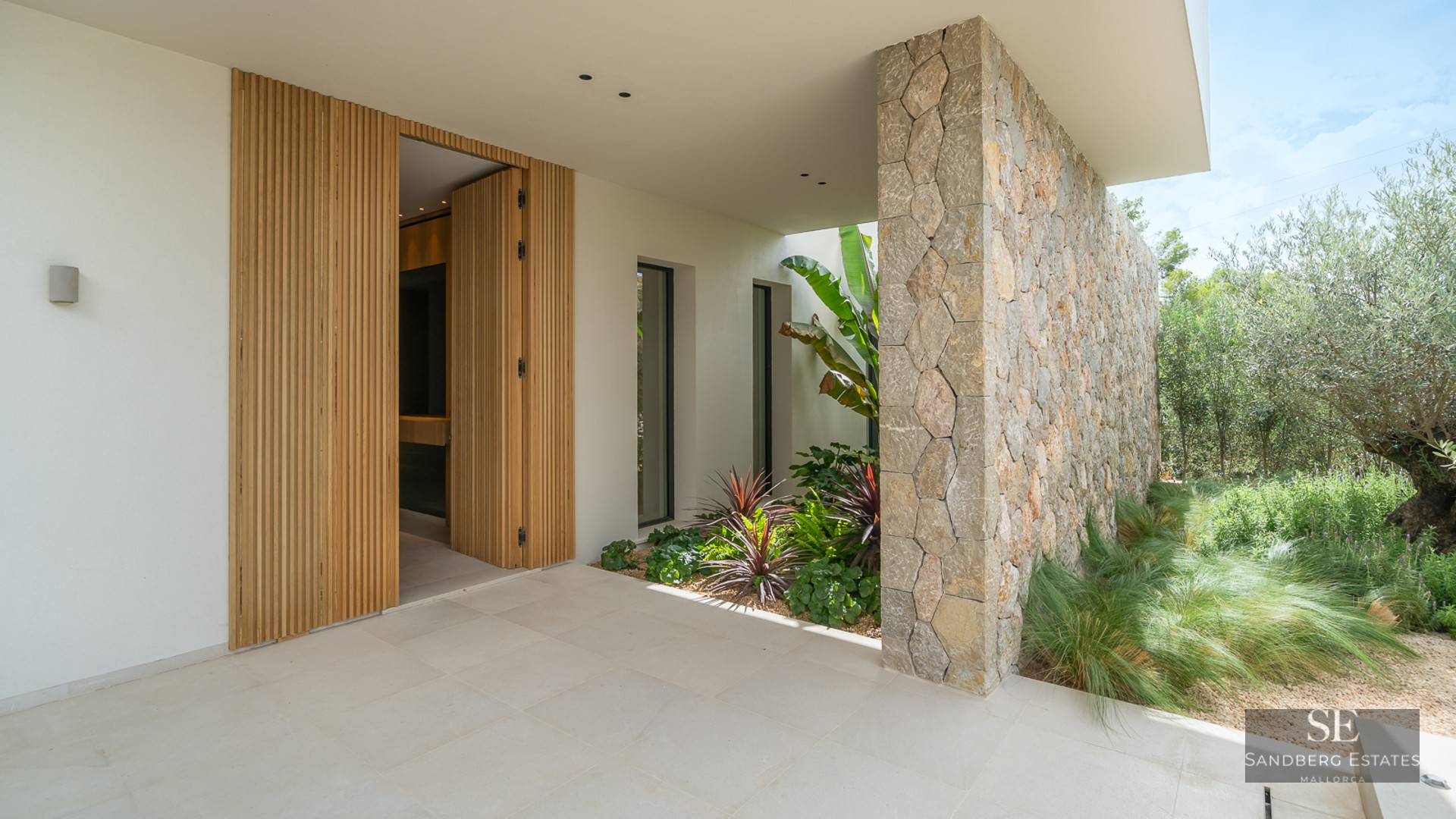 Modern home entrance featuring a slatted wood door, natural stone wall, and lush tropical landscaping.