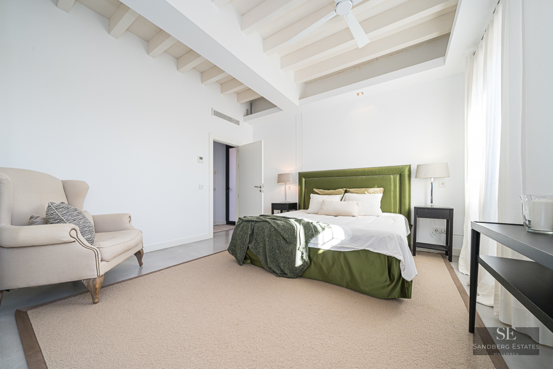 Bright bedroom with white exposed beams, olive green velvet bed, beige wingback chair, and large windows.
