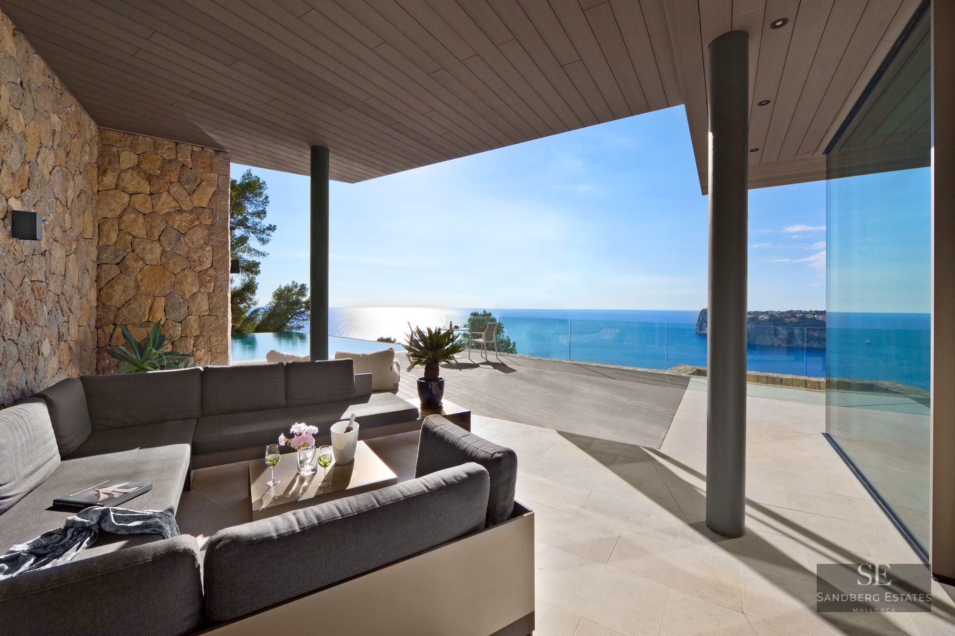Outdoor lounge with grey sofas on a stone terrace overlooking an infinity pool and the Mediterranean Sea.