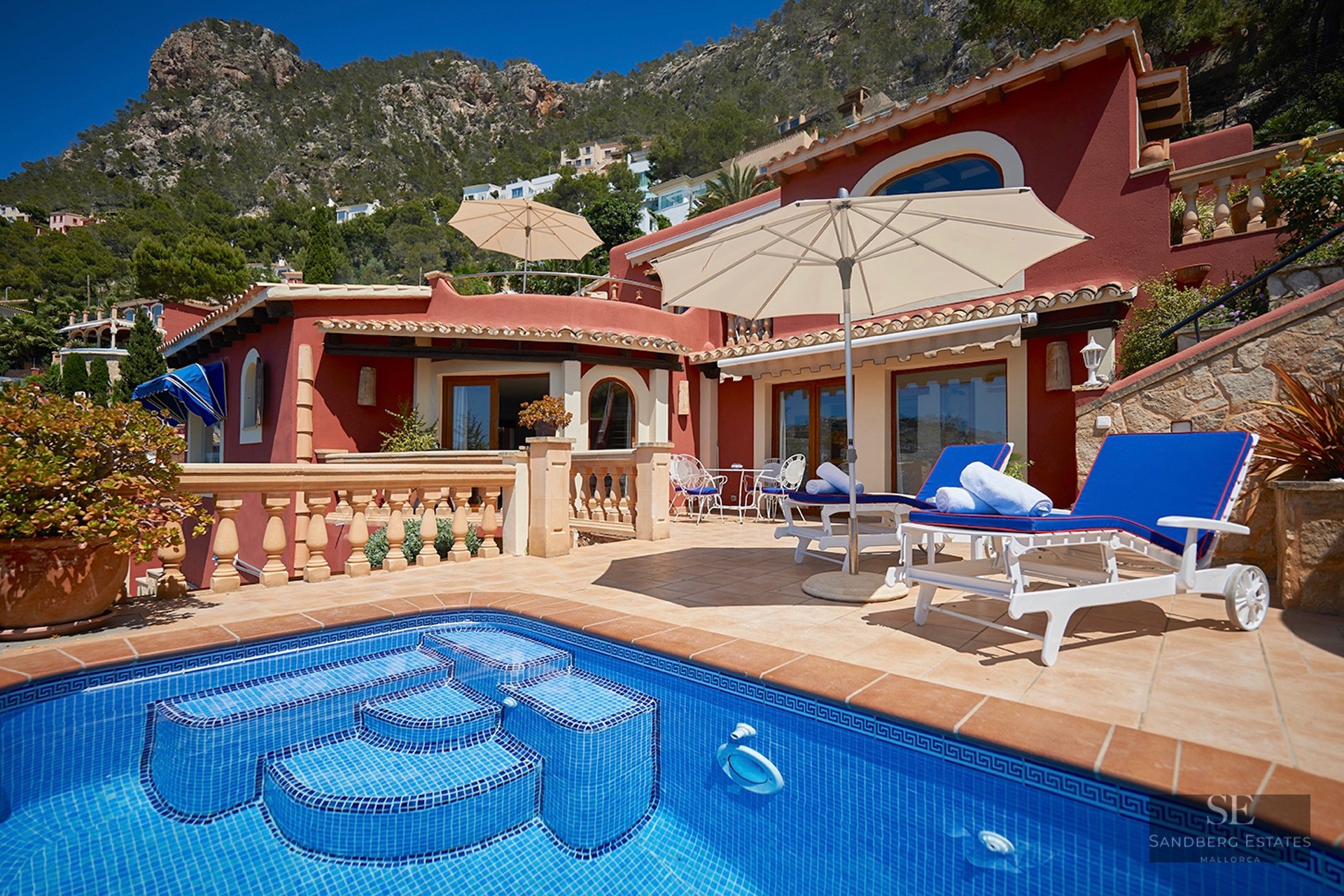 Blue swimming pool and sun deck with loungers in front of a terracotta villa with mountain backdrop.
