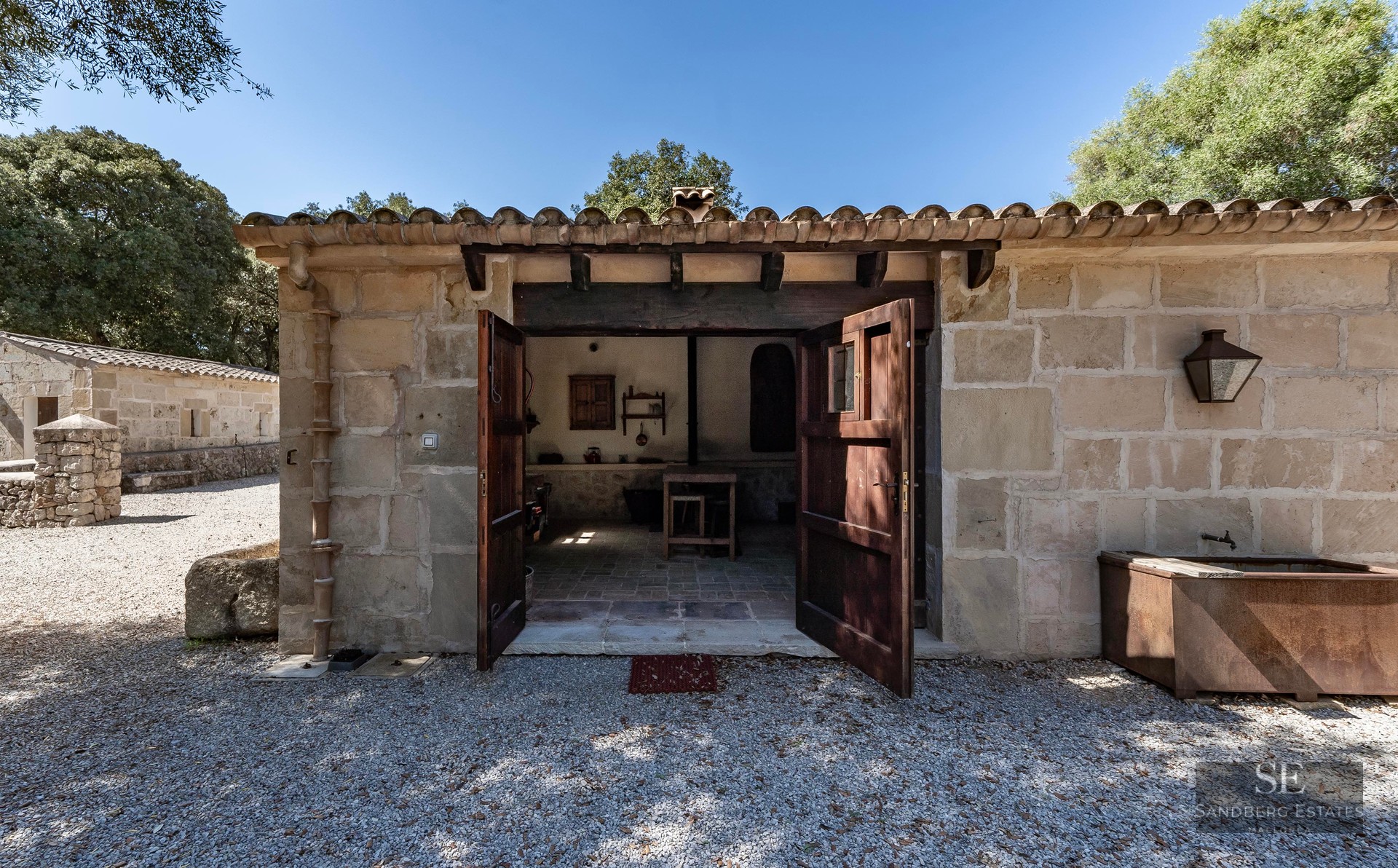 Small rustic stone building with open double wooden doors and a tiled roof on a sunny day.