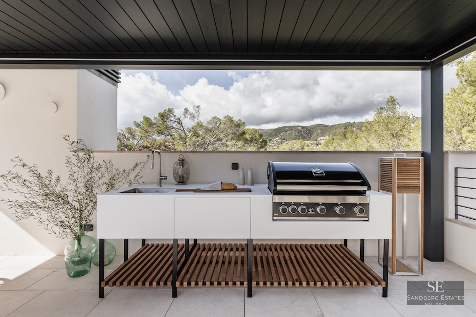 Modern outdoor kitchen with professional BBQ, sink, and white cabinetry on a terrace overlooking a mountain landscape.