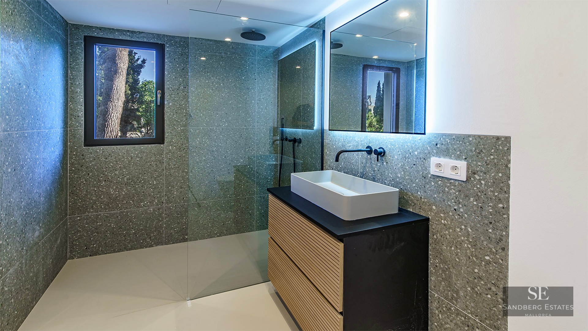 Modern bathroom with grey terrazzo walls, a walk-in glass shower, black fixtures, and a wooden vanity with a white sink.