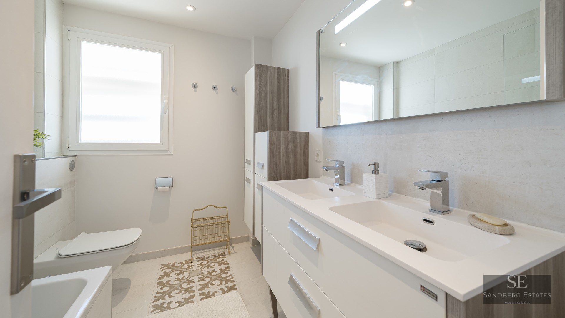 Bright modern bathroom with a double white vanity, large mirror, wall-hung toilet, and natural window light.