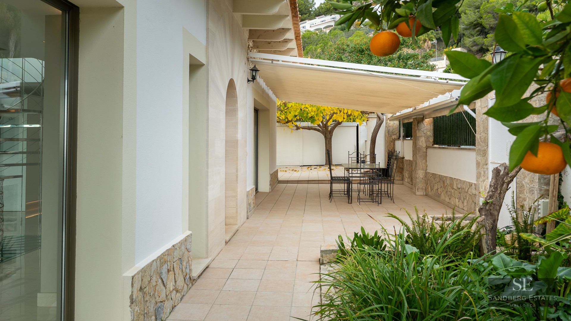 A sun-shaded outdoor terrace with a dining table, black metal chairs, stone walls, and a lush orange tree.