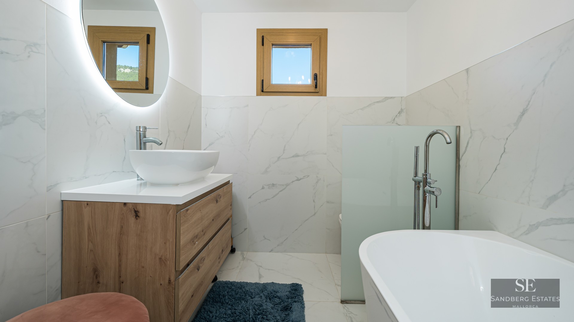 Modern bathroom with marble walls, wood vanity, vessel sink, circular backlit mirror, and a white bathtub.