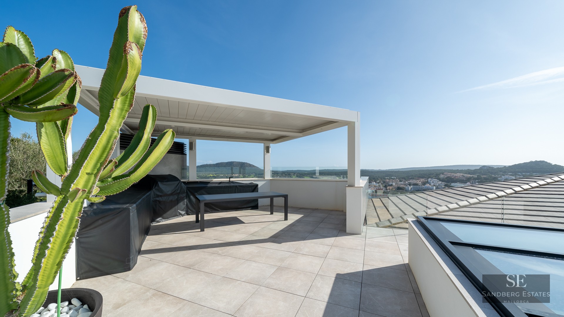 Modern rooftop terrace with a white pergola, outdoor kitchen, large cactus, and scenic views under a clear sky.