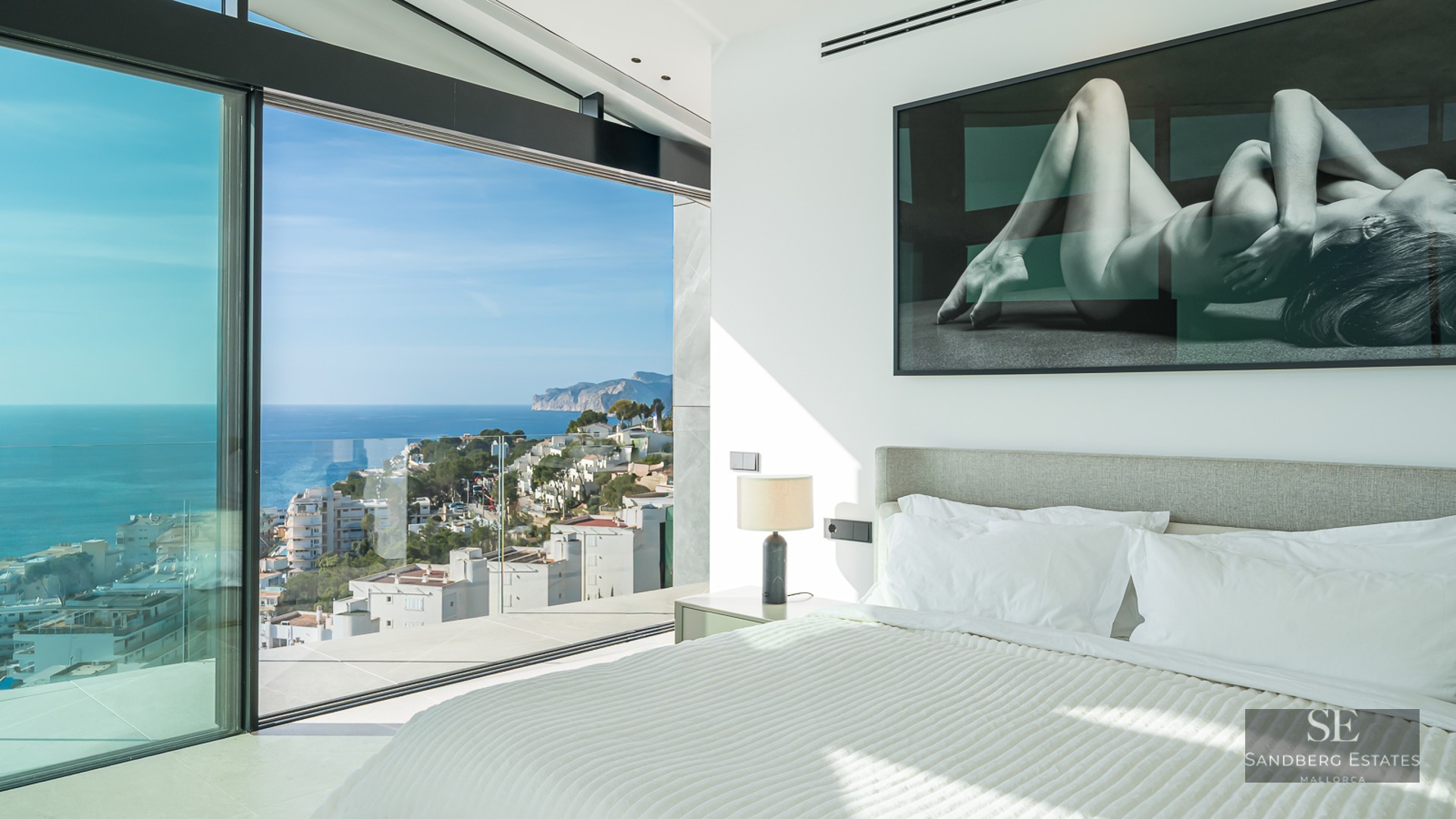 Modern bedroom with large glass windows showing a panoramic view of the sea and coastline.