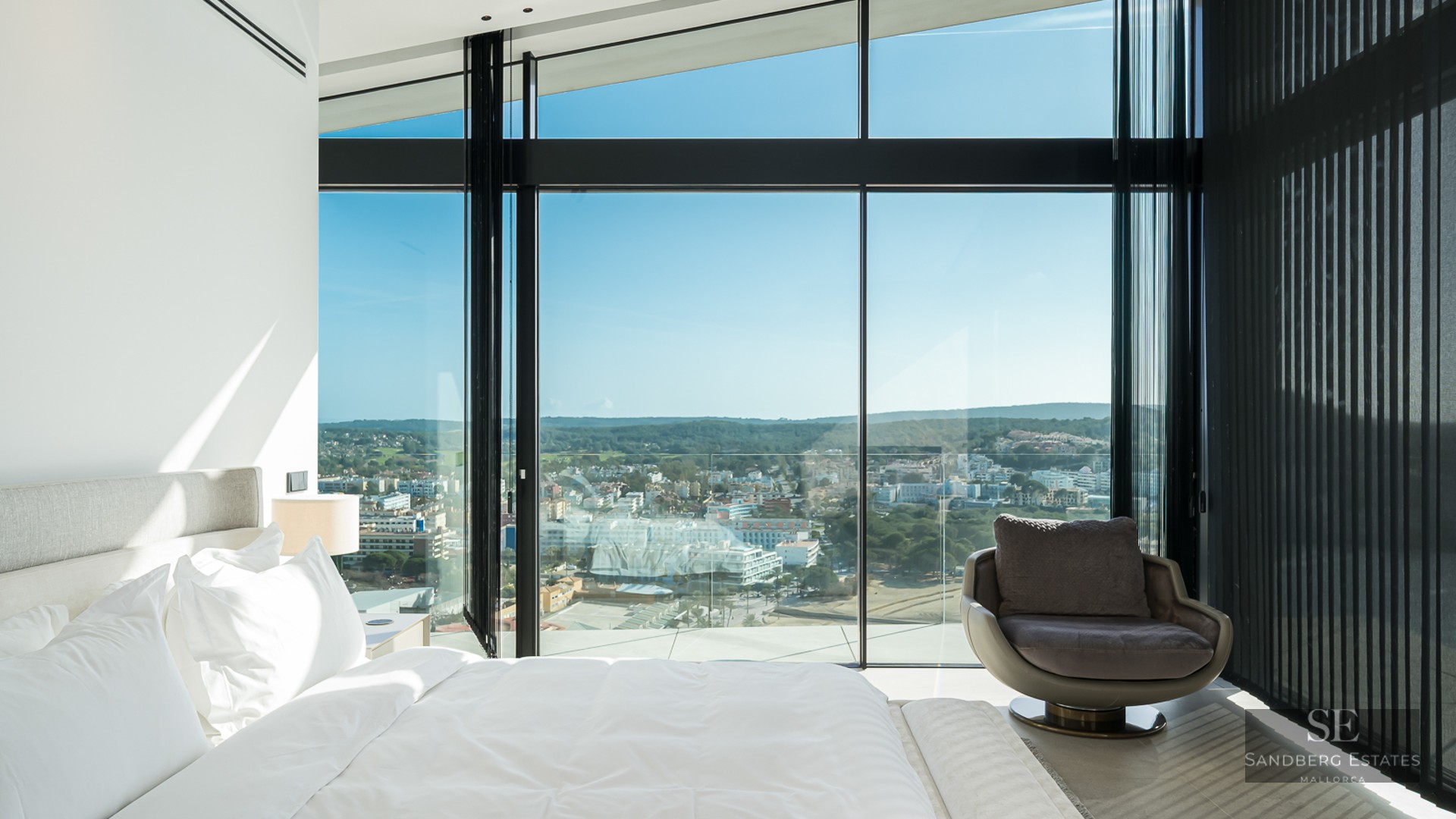 Modern master bedroom featuring floor-to-ceiling windows with a sweeping city view and a designer armchair.