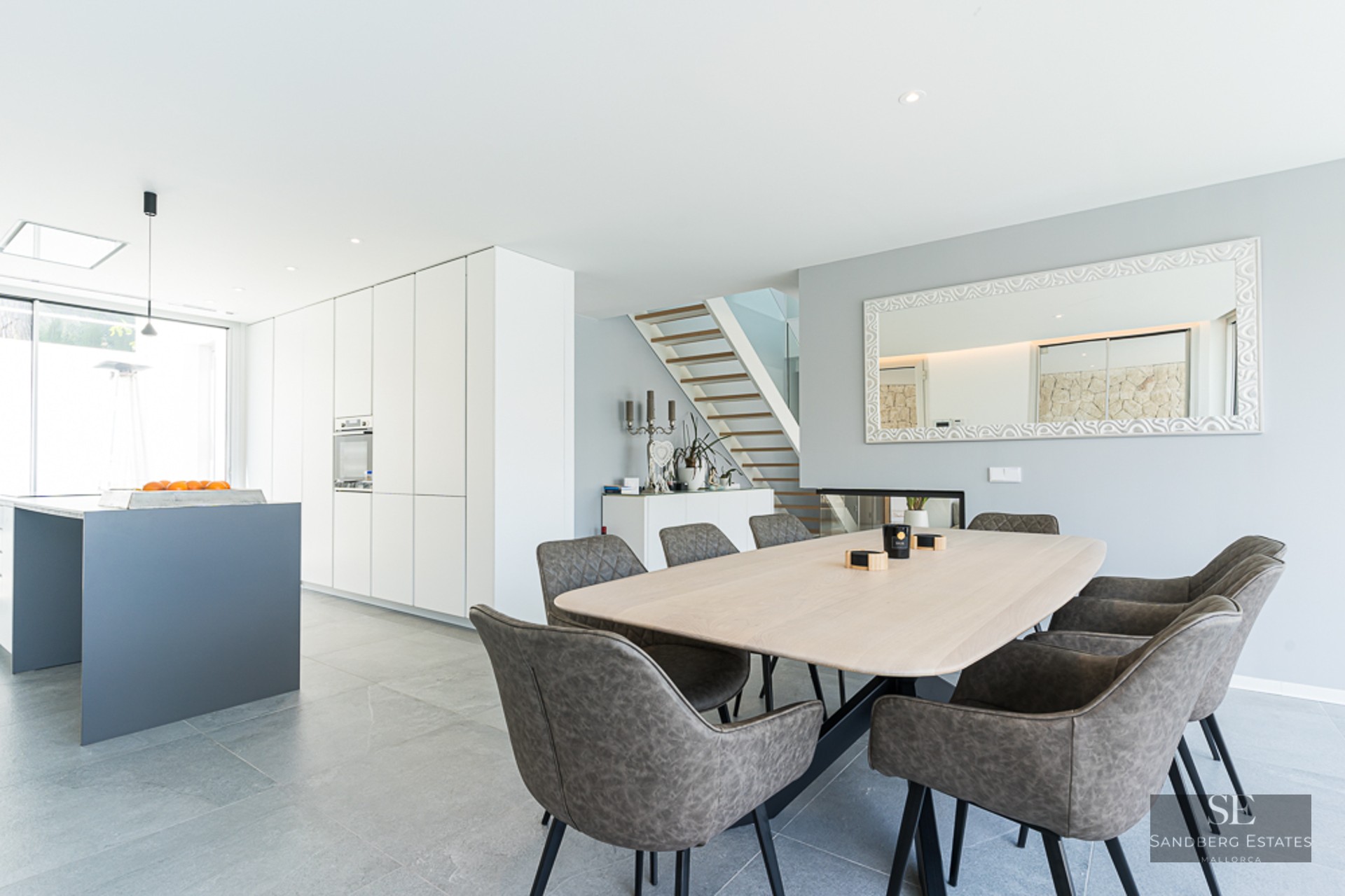 Bright dining area featuring a light wood table, grey chairs, and a minimalist white kitchen in the background.