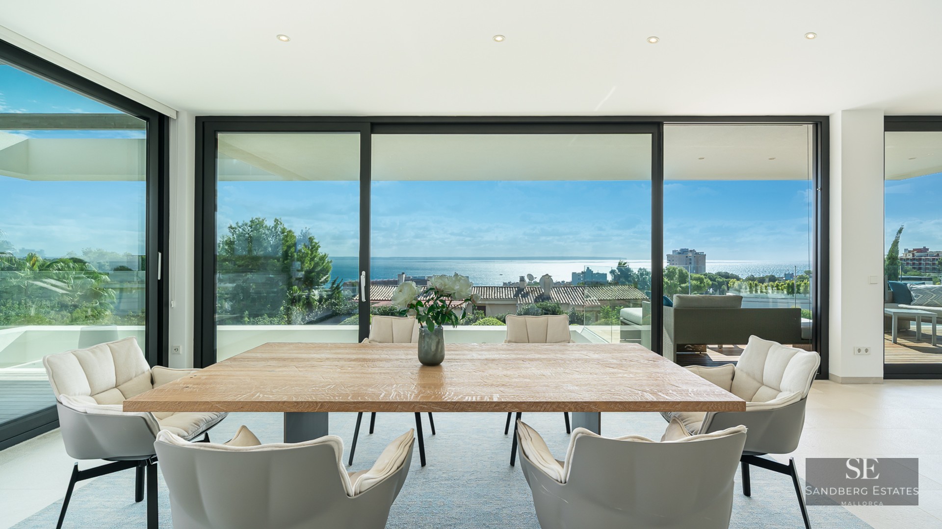 Bright dining room with a large wooden table and grey chairs facing floor-to-ceiling windows with a vast ocean view.