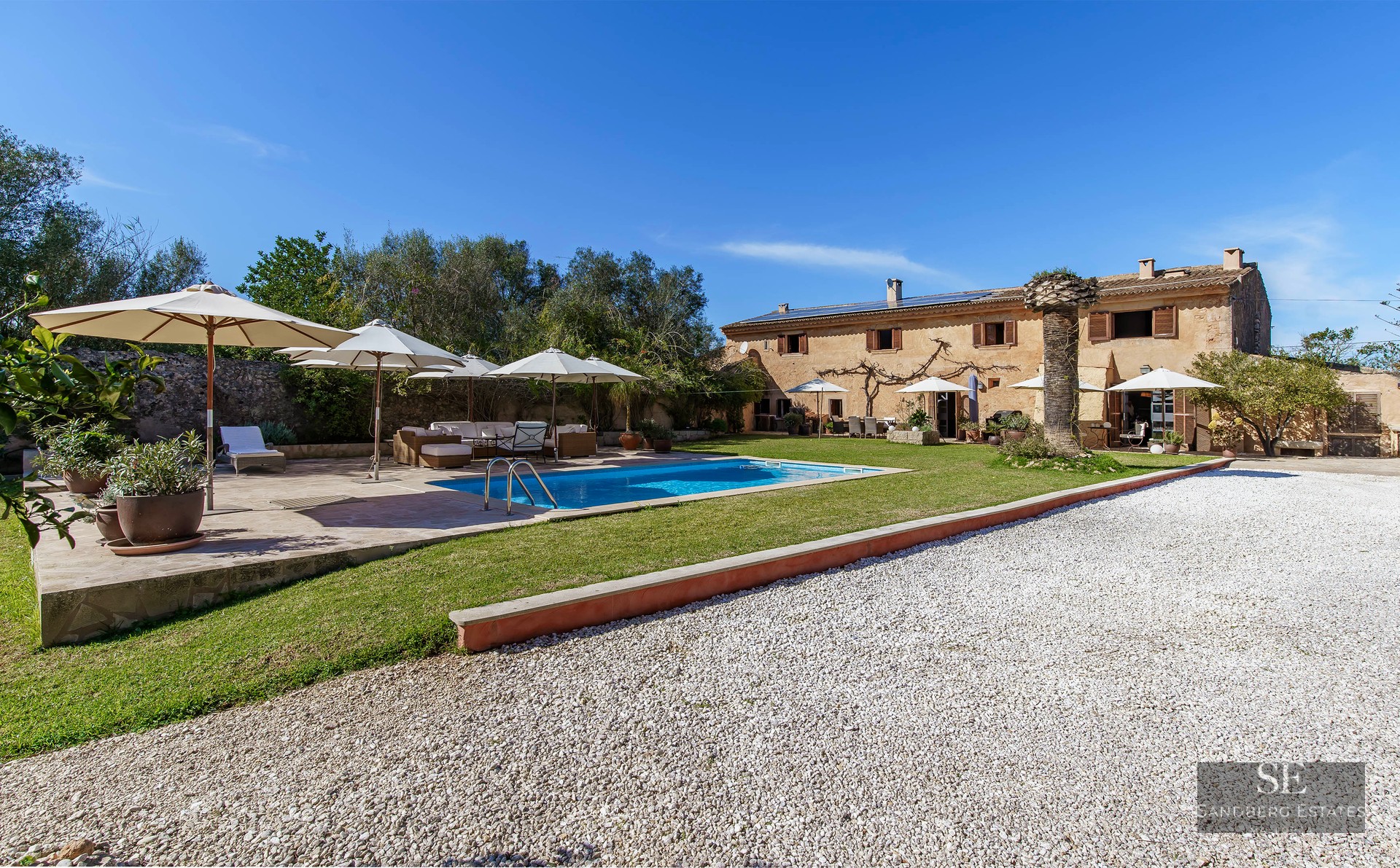 View of a large stone villa with a blue swimming pool, white umbrellas, and garden under a clear sky.