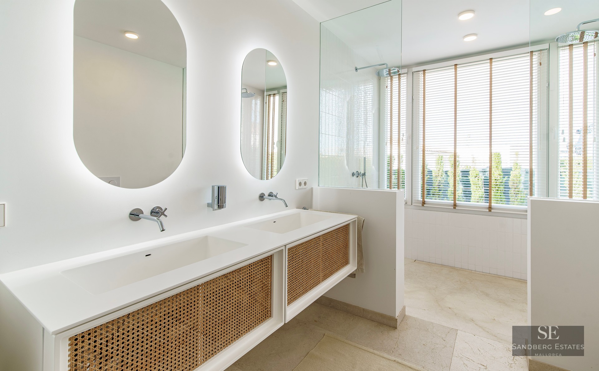 Modern master bathroom featuring a white double vanity with rattan doors, oval backlit mirrors, and a glass-enclosed shower.