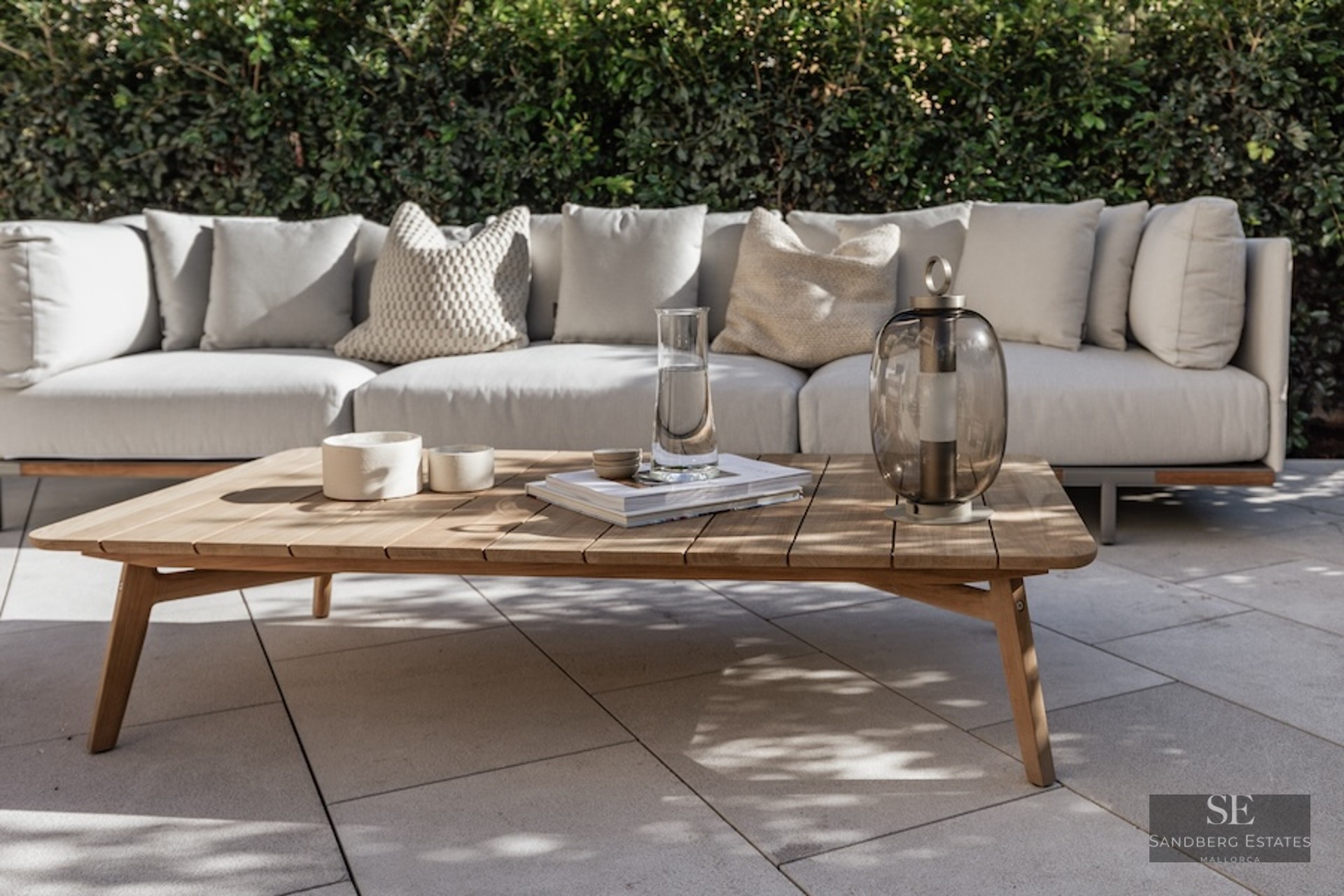 Outdoor sofa with beige cushions and a wooden coffee table on a stone patio against a green hedge.