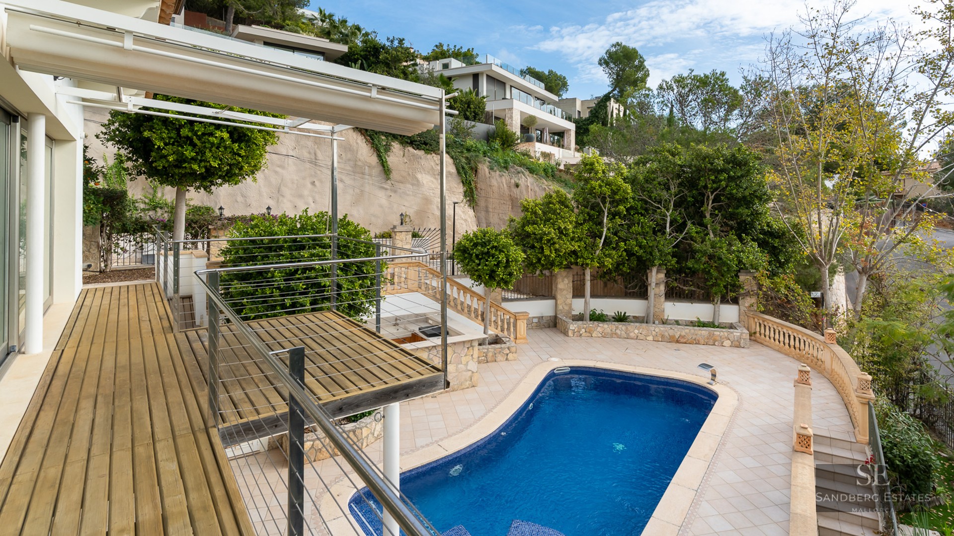 High-angle view of a blue swimming pool, wooden deck, and tiled patio set against a lush green hillside.