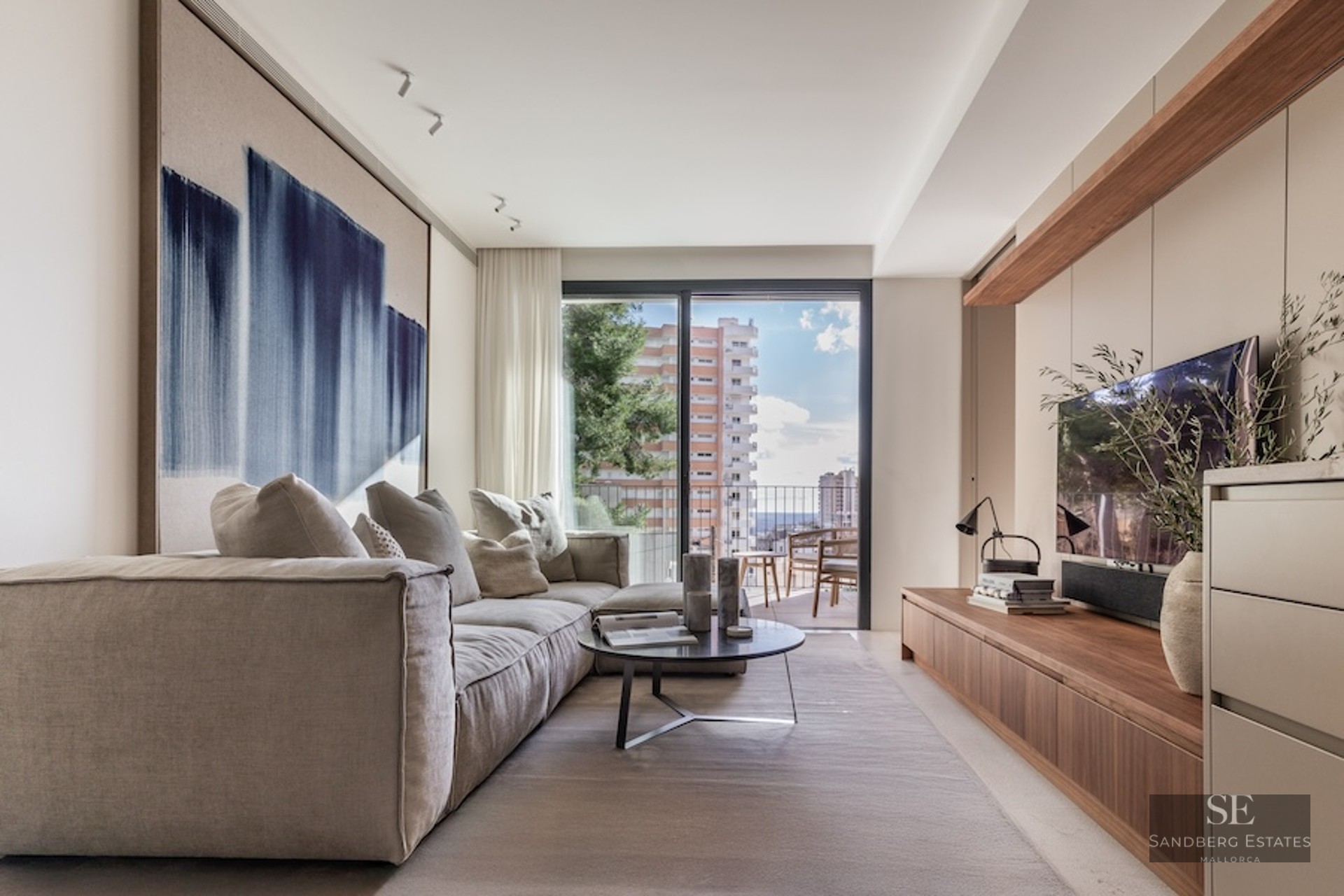 Bright modern living room featuring a plush beige sofa, abstract blue art, wooden TV console, and a balcony view.
