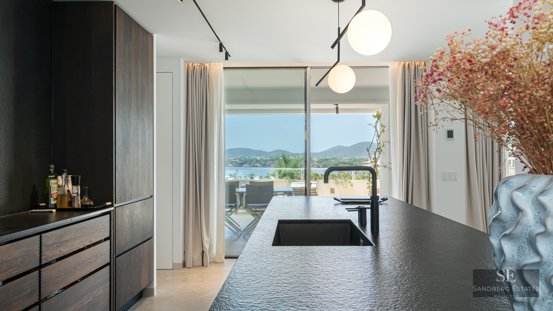 Modern kitchen with a dark stone island and black sink looking out to a sunlit terrace and sea view.