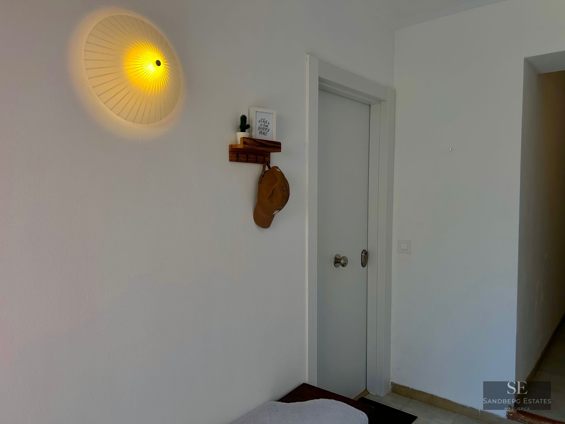 A white hallway featuring a round pleated wall lamp, a small wooden shelf with a hat, and a white door.