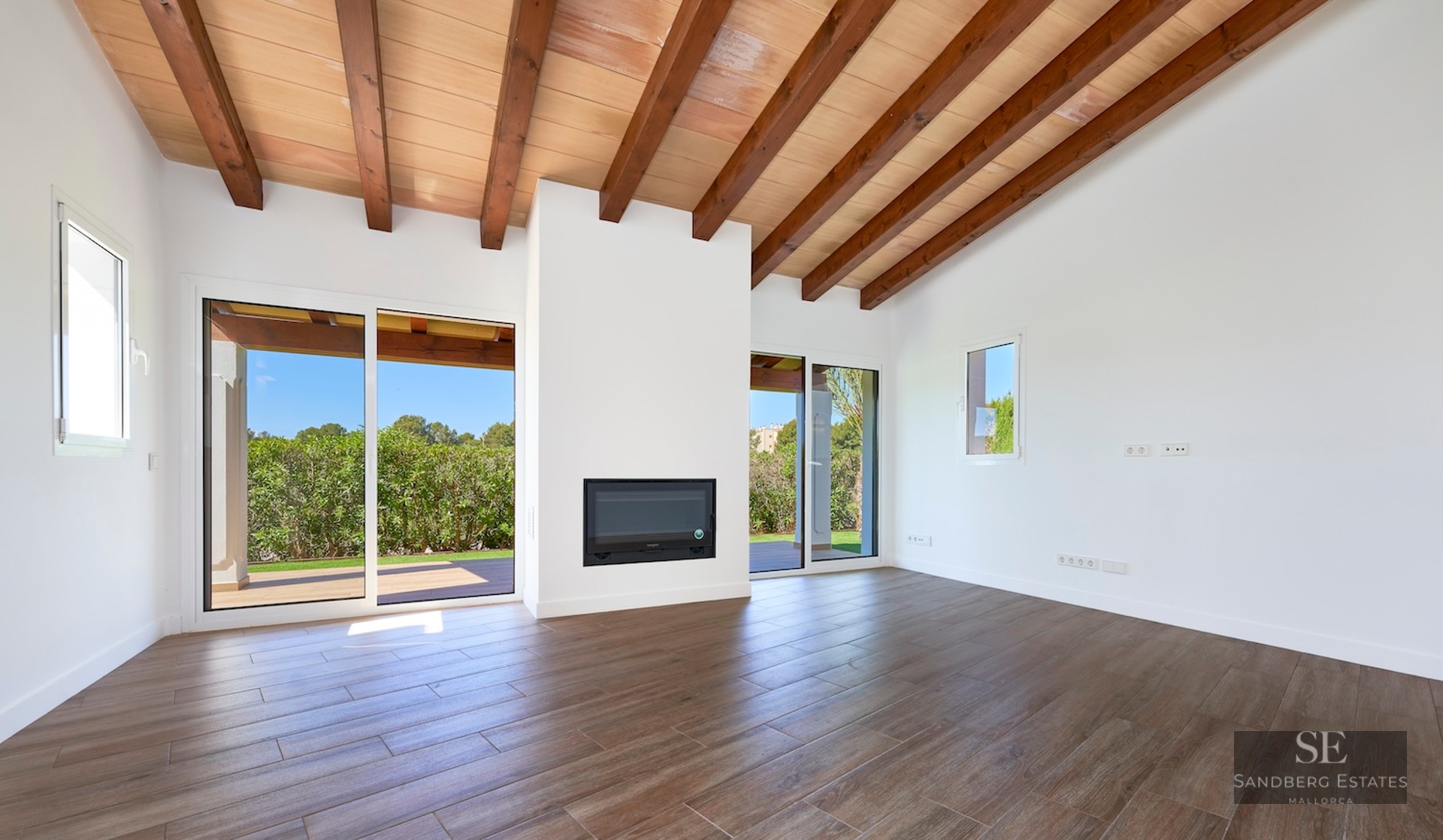 Spacious white living room with high ceiling, exposed wooden beams, fireplace, and sliding glass doors to a garden.