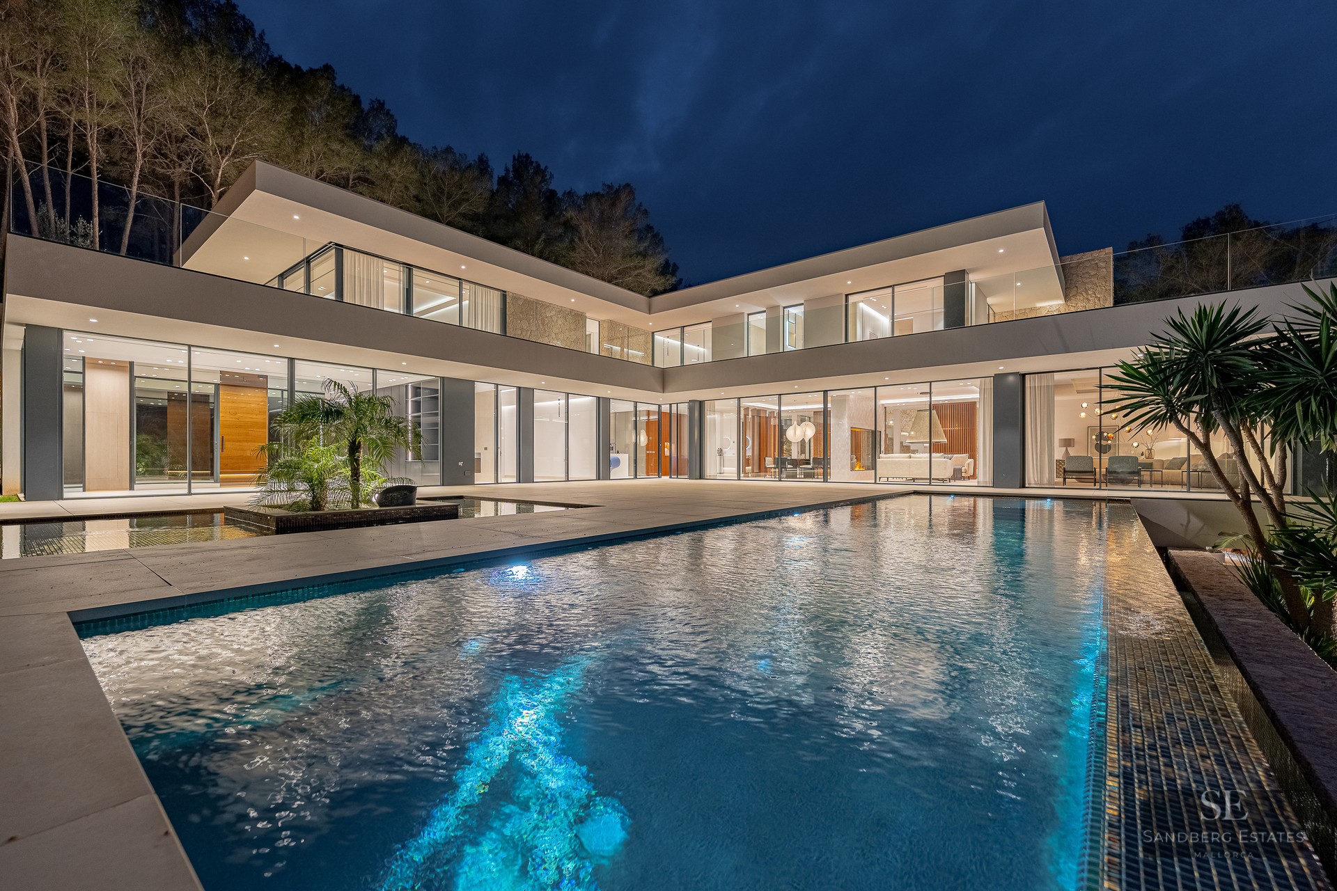 A luxury swimming pool in front of a glowing modern glass villa under a dark night sky.