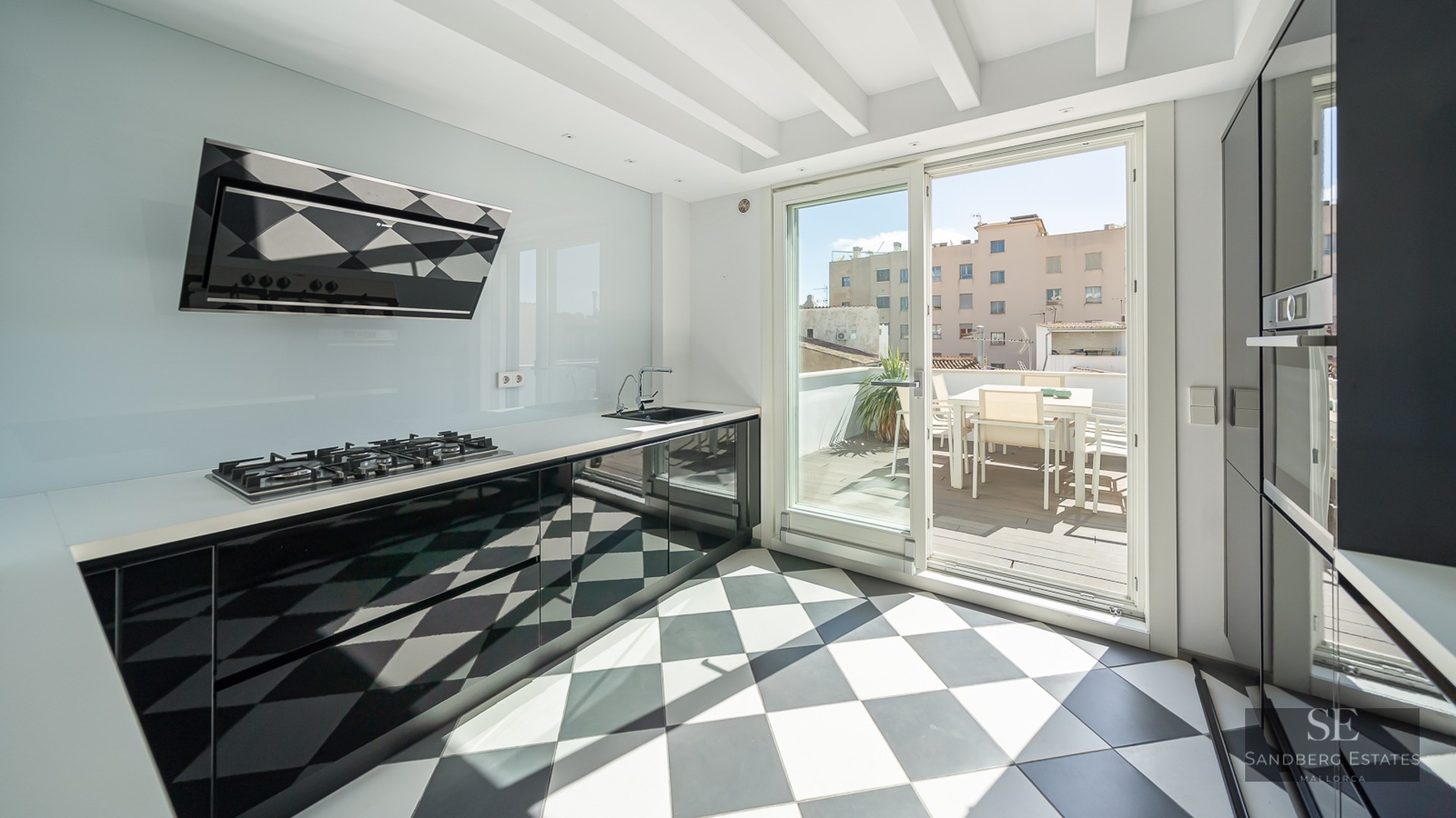 Modern kitchen with black and white checkered floor, glossy black cabinets, and a glass door leading to a sunny terrace.