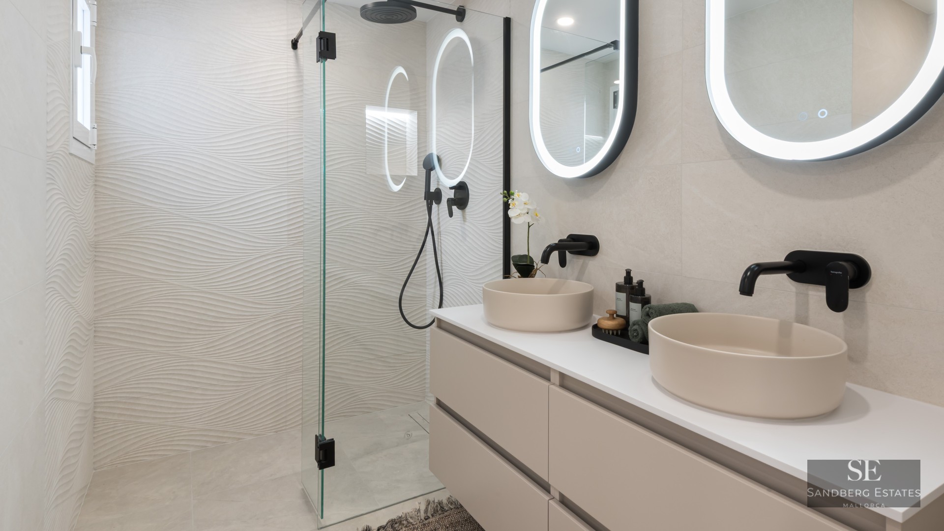 Modern bathroom with two beige vessel sinks, backlit oval mirrors, and a glass shower with textured wavy tiles.