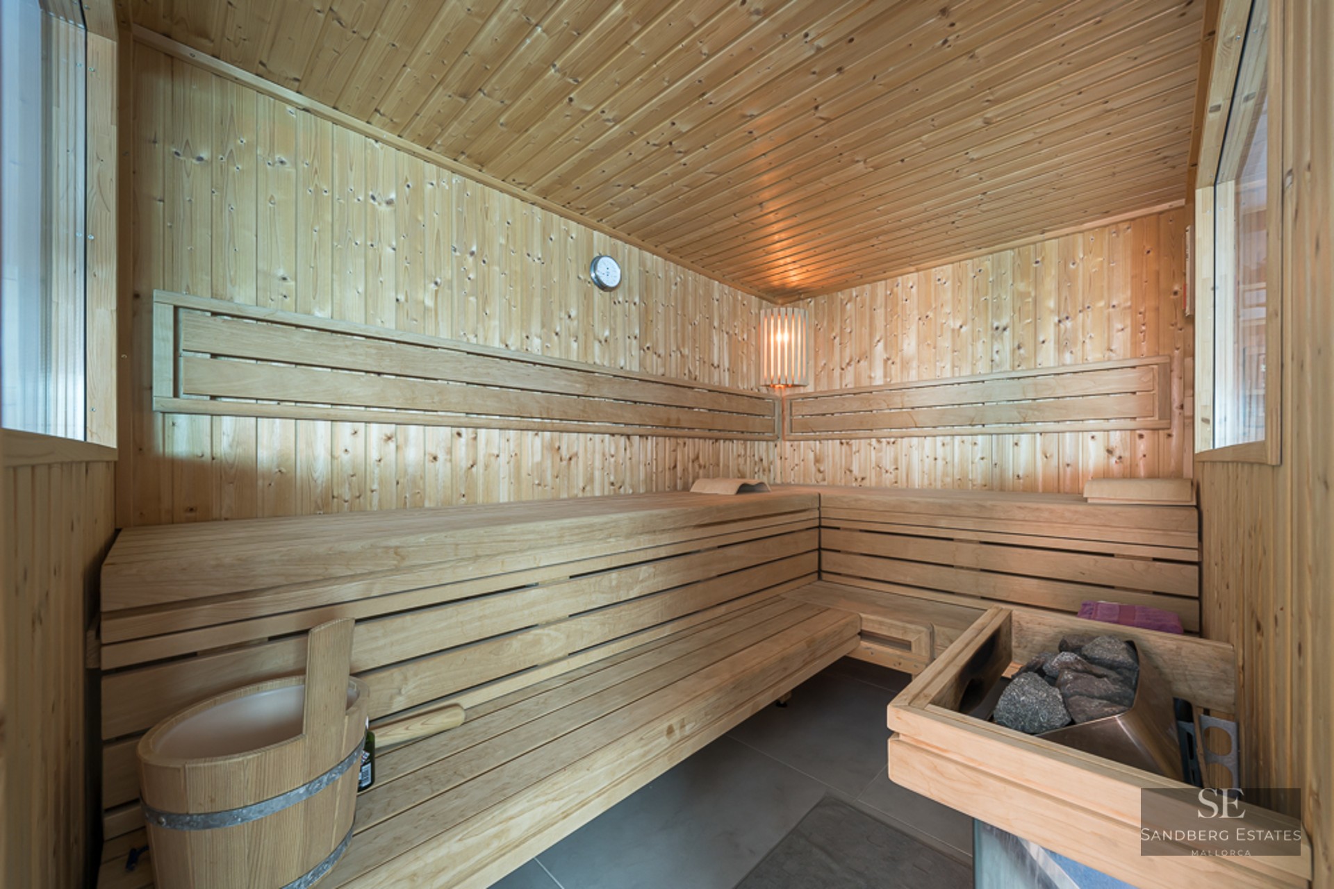Interior of a modern wood-paneled sauna with tiered seating, a stone heater, and a wooden bucket.