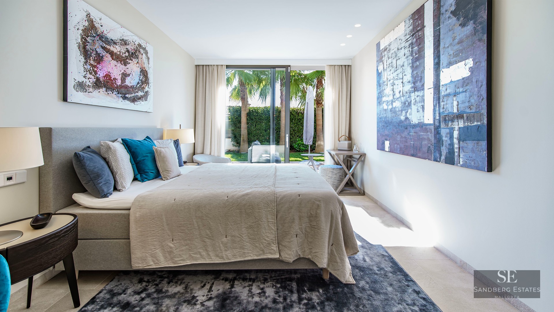 Chambre moderne avec grand lit, art abstrait et baies vitrées ouvrant sur un jardin avec des palmiers.